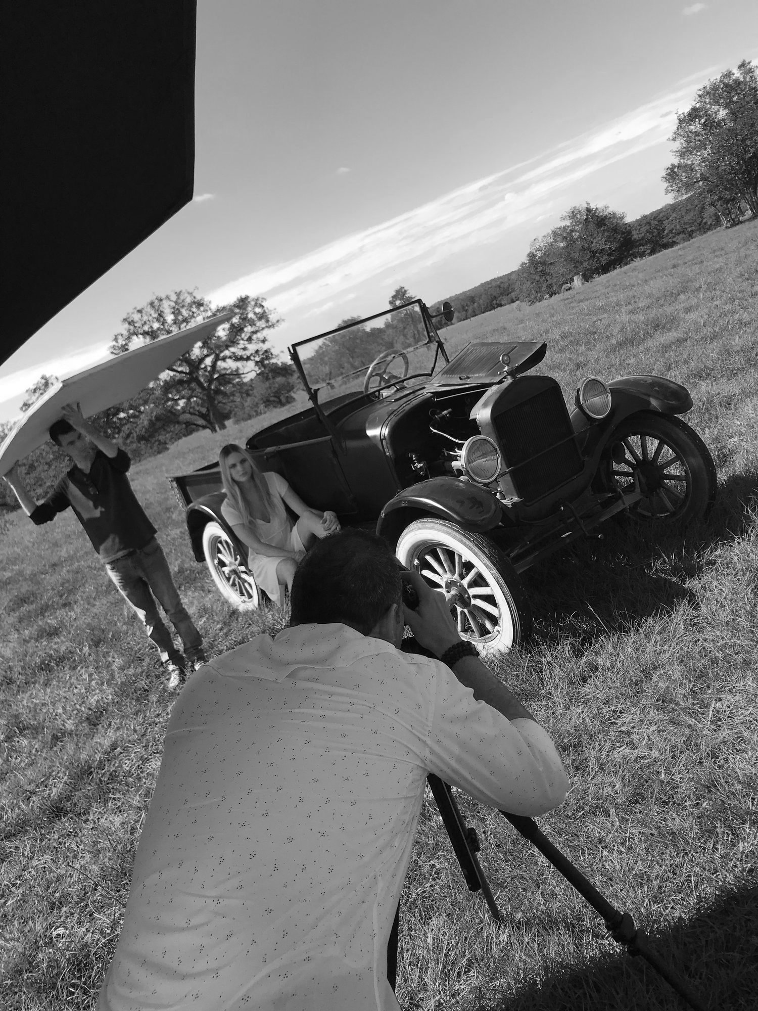 A photoshoot scene with a vintage car on a grassy field, a woman sitting beside the car, a man with a camera taking photos, and another person holding a reflector in the background.