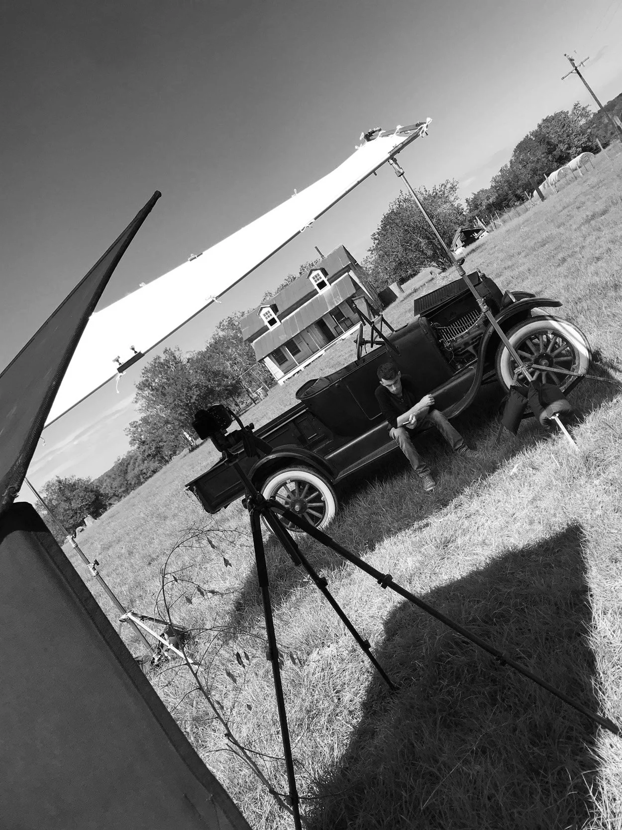 A black-and-white photo of a vintage car set up as a filming or photo shoot scene outdoors. A man is sitting on the side of the car, looking at his phone. There are large studio lights and reflectors set up around the car, with a building and trees in the background.