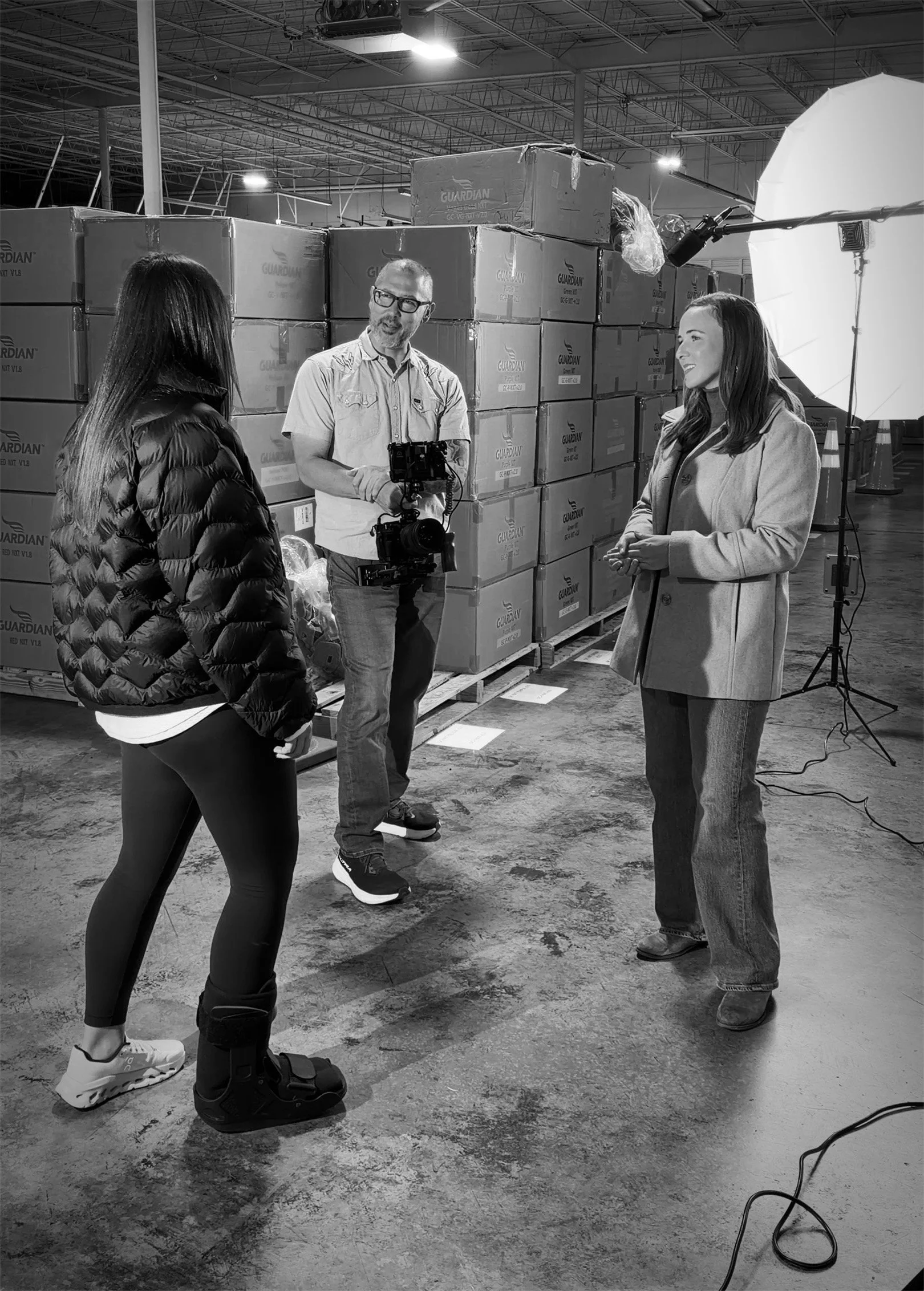 A photo of three people in a warehouse, filming a segment. Two women and one man are present, with the woman on the left in a black jacket, the man in the middle with glasses holding a camera, and the woman on the right speaking. Large boxes labeled 'Guardian' are stacked behind them, and professional lighting and equipment are set up.