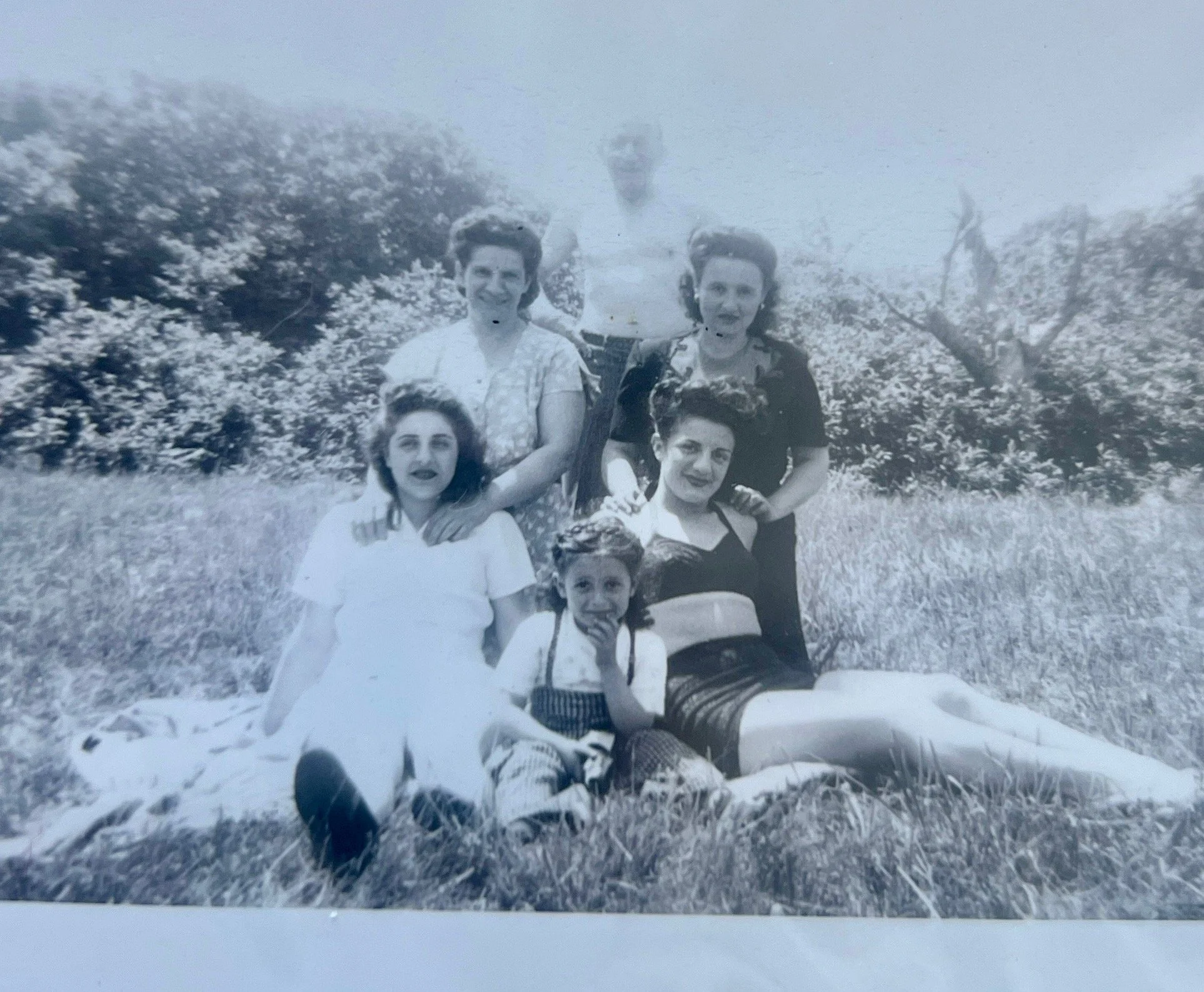 Black and white photograph of seven women and children sitting and standing on grass outdoors, with trees in the background.