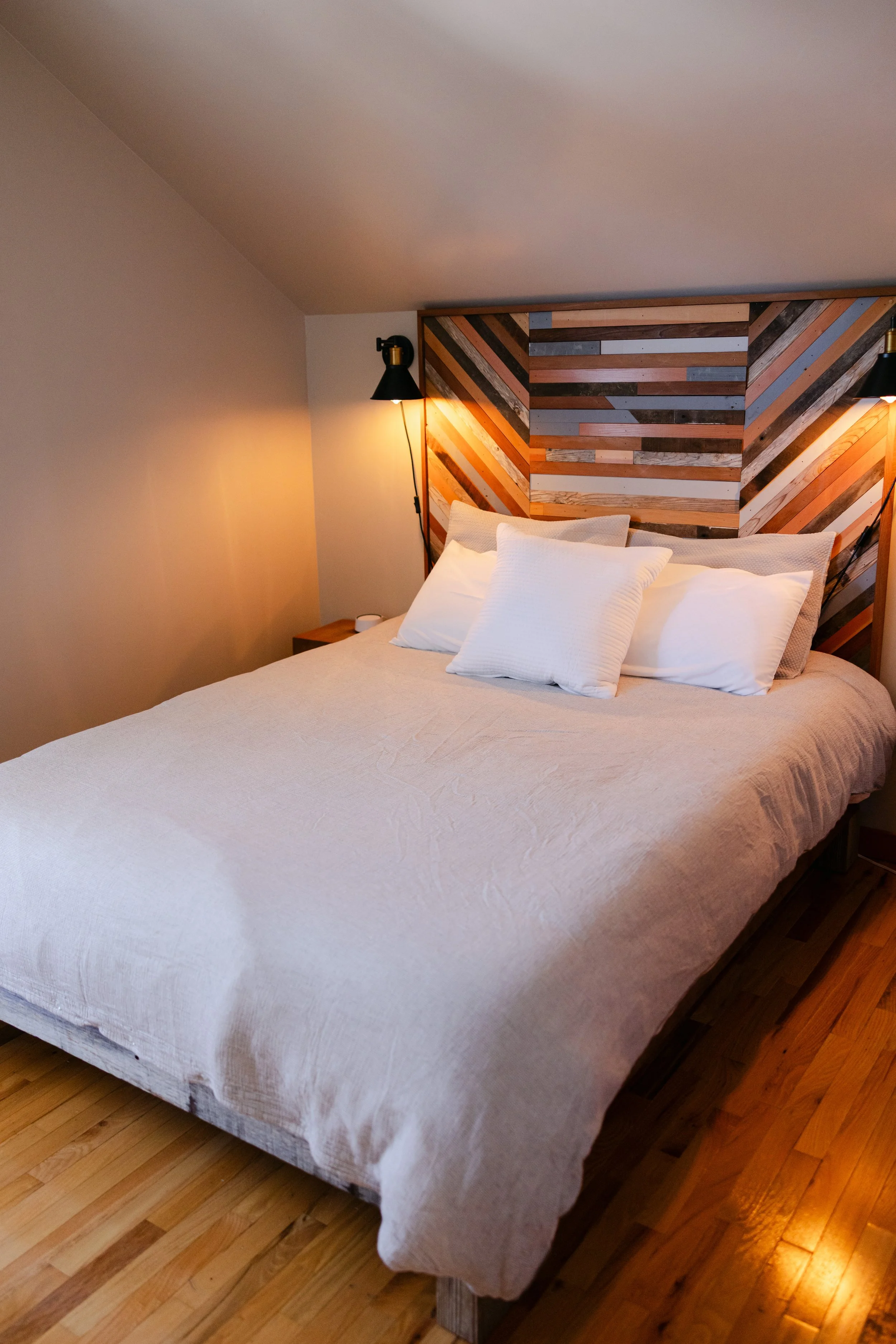 A neatly made bed with white bedding and pillows, positioned against a wooden wall with decorative diagonal wood paneling, in a bedroom with hardwood floors and wall-mounted lamps.