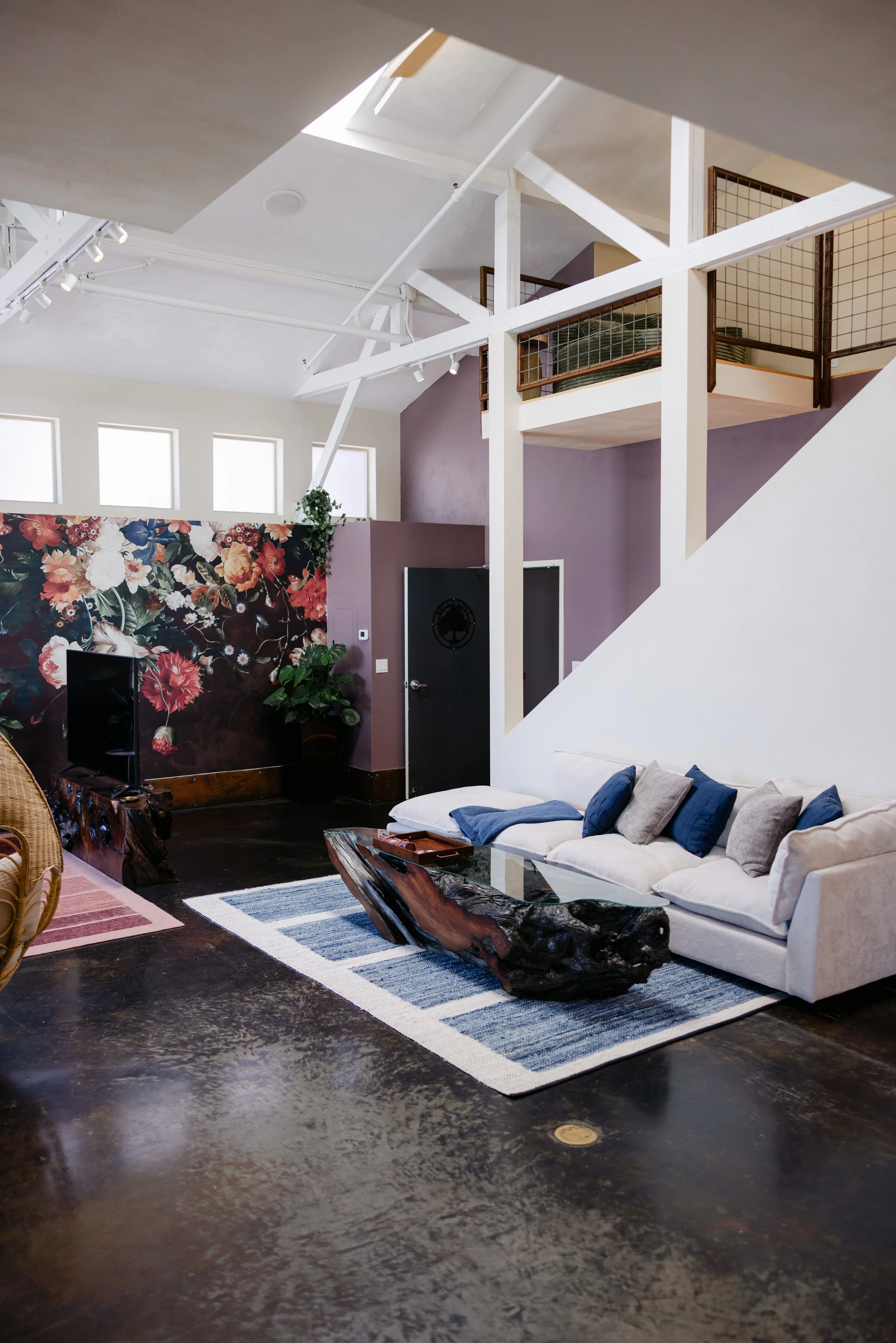 Modern living room with white sofa, blue and gray pillows, wooden coffee table, floral wallpaper, and purple accent wall. Open loft area with railing, skylight, and large windows.