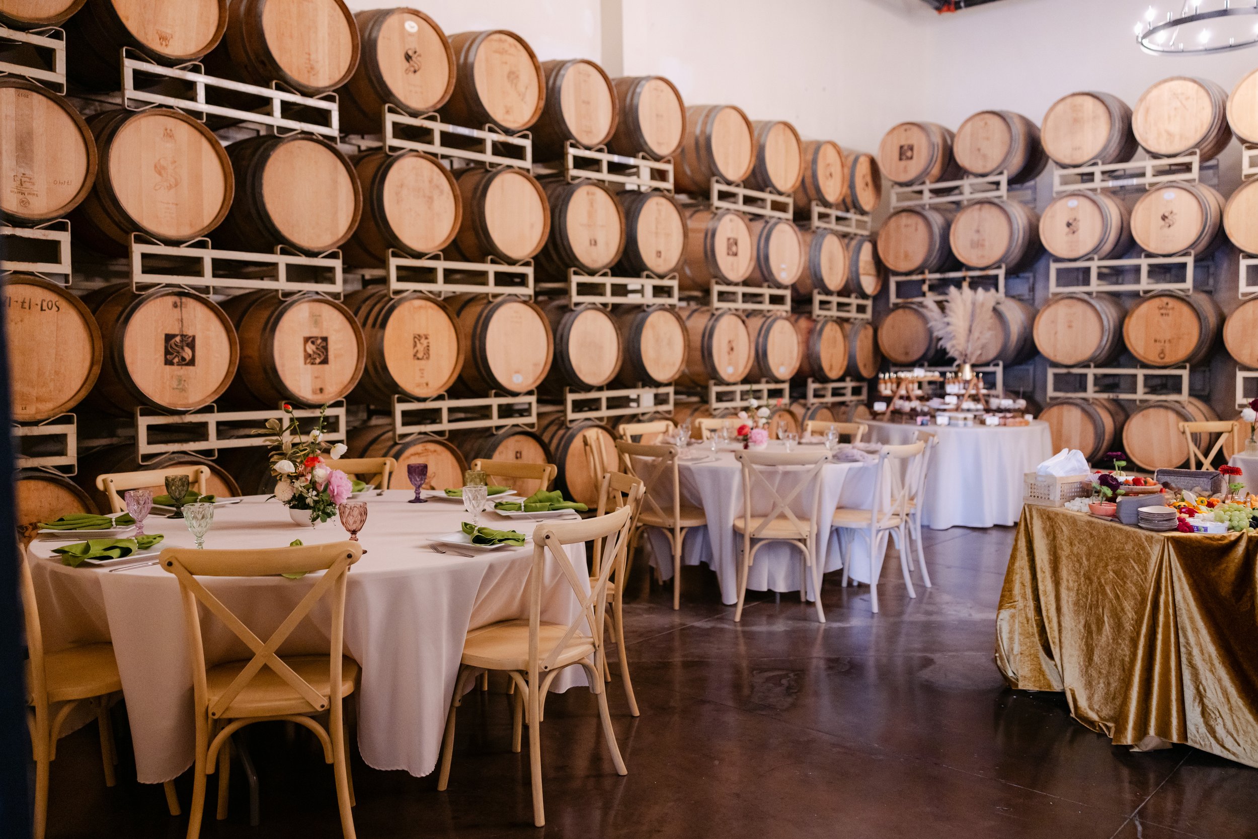 Interior of a winery or wine cellar with wooden wine barrels stacked on shelves, and a dining area with tables set with green napkins, floral centerpieces, and glasses, indicating a celebration or event.