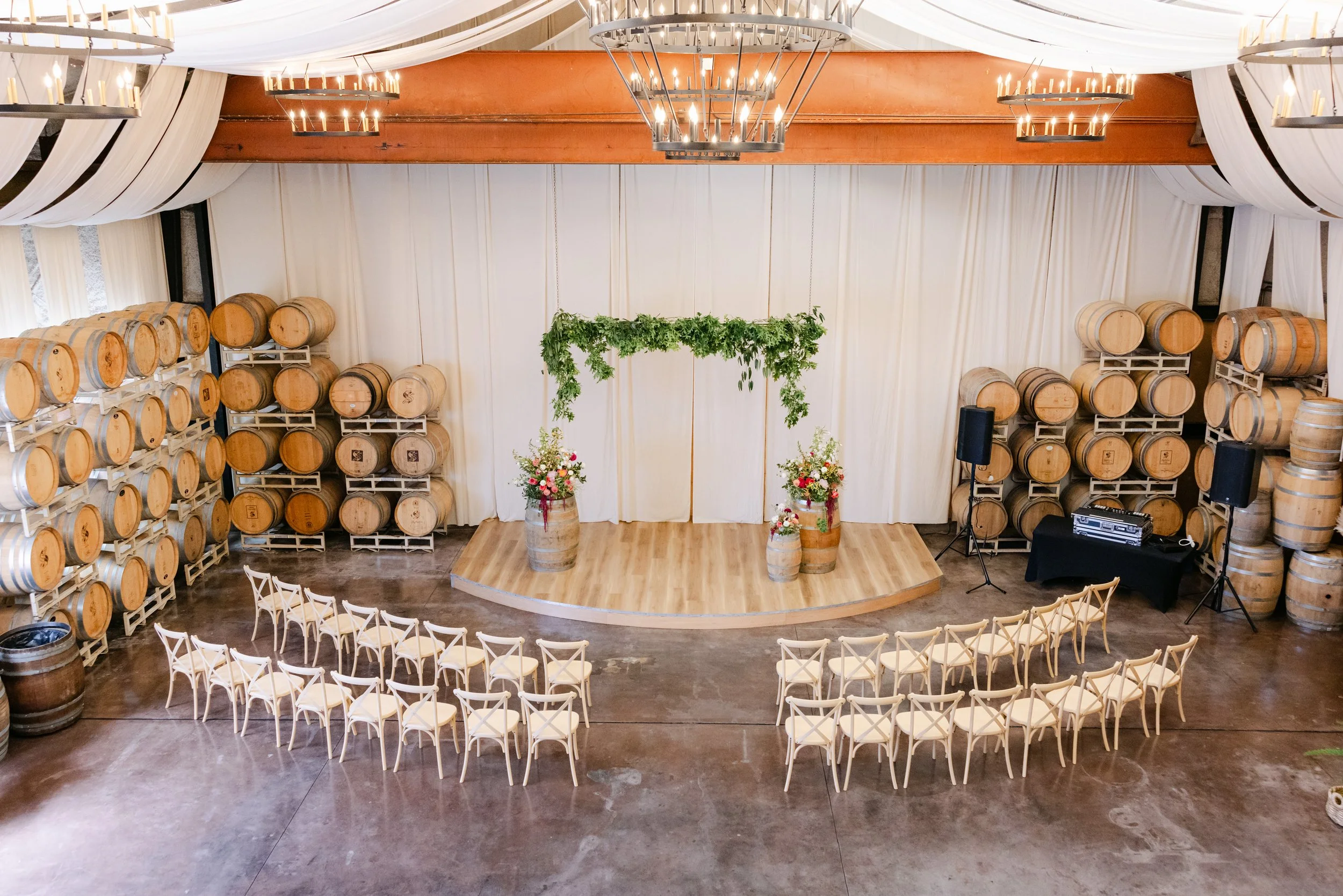 An indoor wedding venue decorated with white drapes, chandeliers, and wooden barrels. The ceremony space has rows of white chairs facing a small stage with floral arrangements and greenery hanging above.