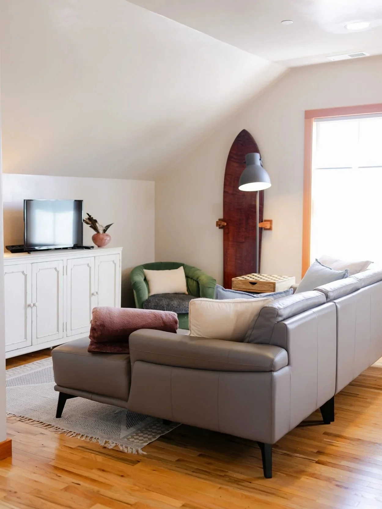 Living room with gray sofa, green armchair, white cabinet with TV, decorative vase, and surfboard wall art, hardwood floors, and window with natural light.