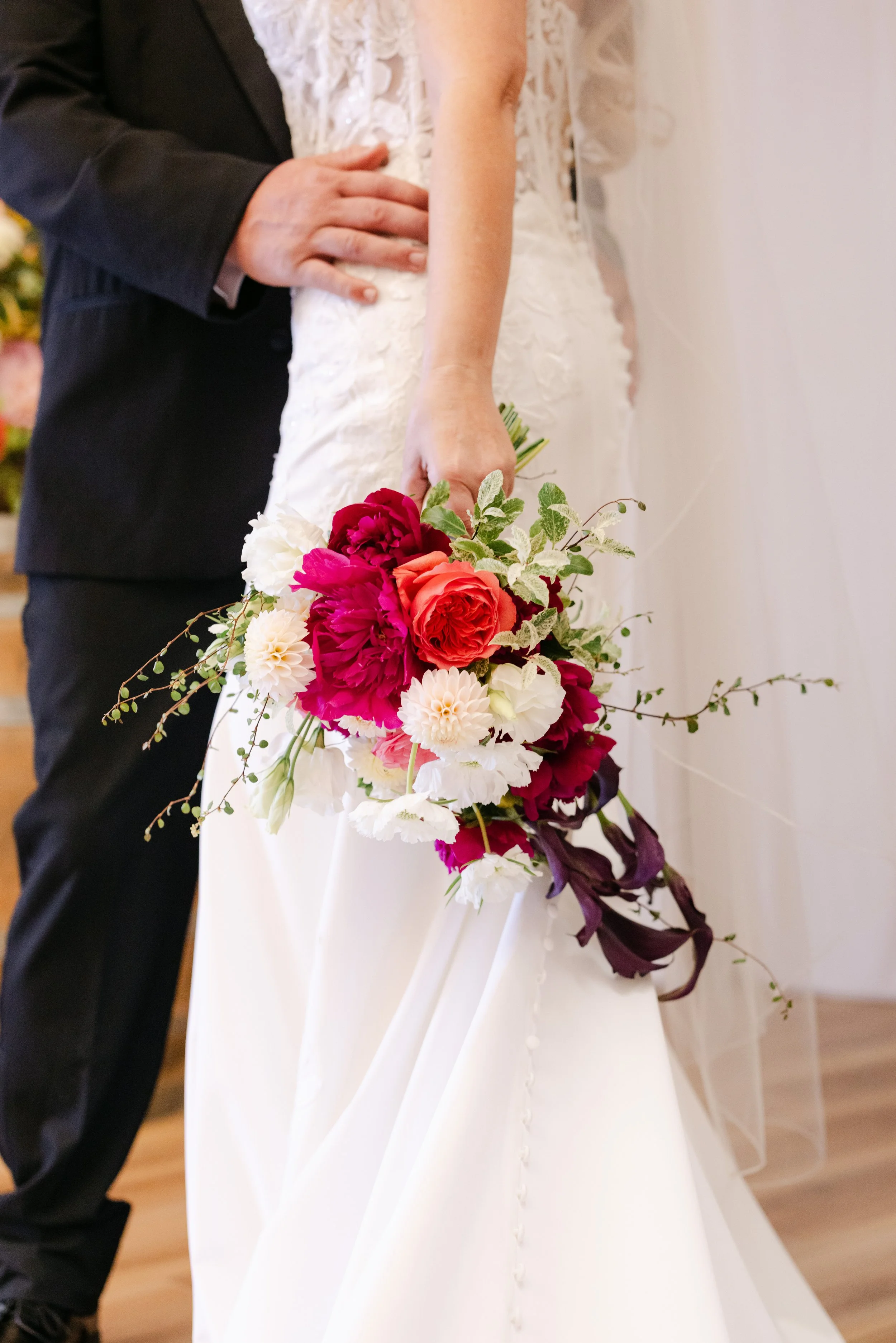 A bride in a white wedding gown holding a vibrant bouquet with red, pink, white, and purple flowers, and a groom in a black tuxedo standing next to her.