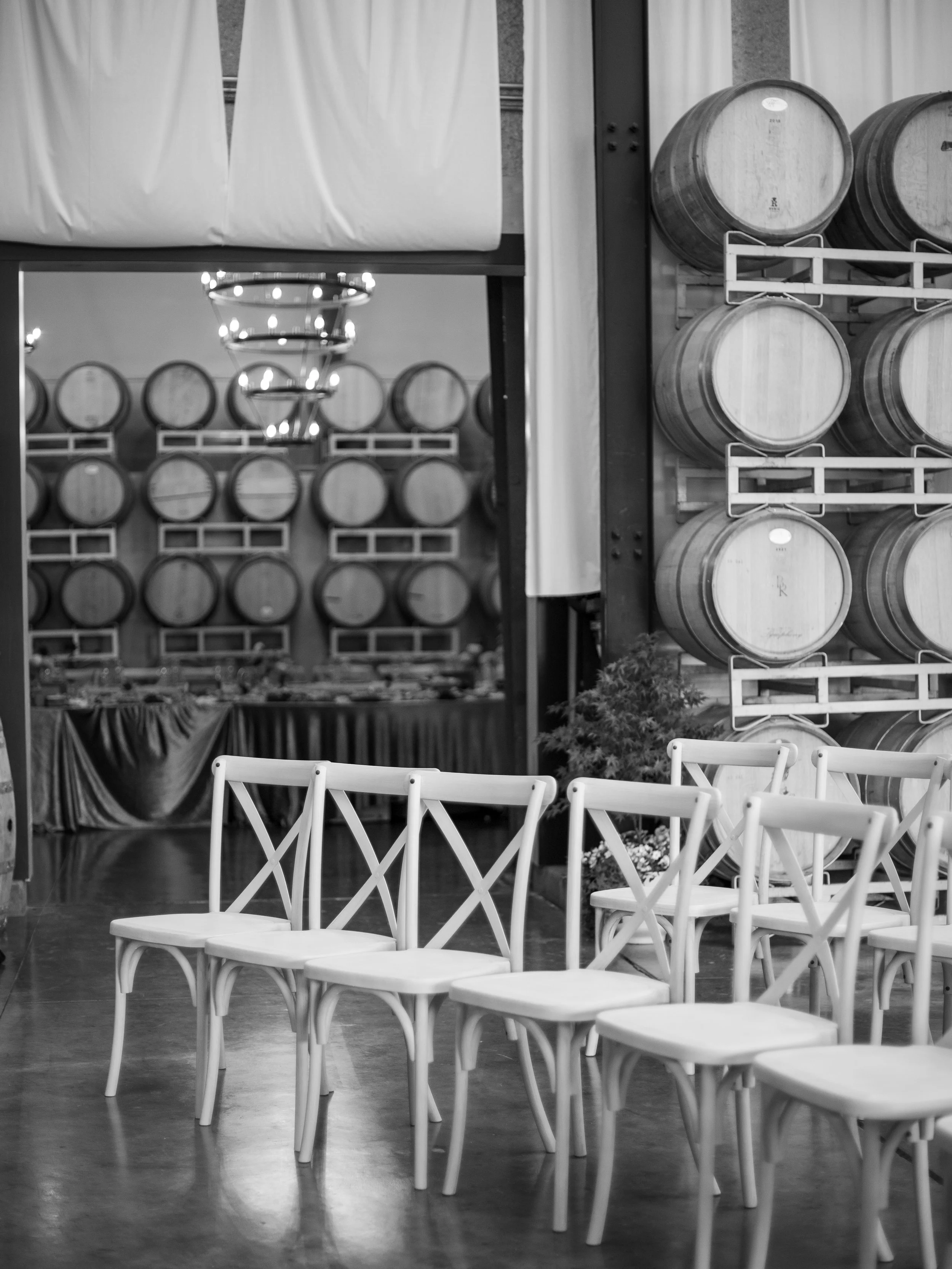 Black and white photo of a venue with white chairs arranged in rows, with some barrels stacked on stands in the background, likely at a winery or event space.