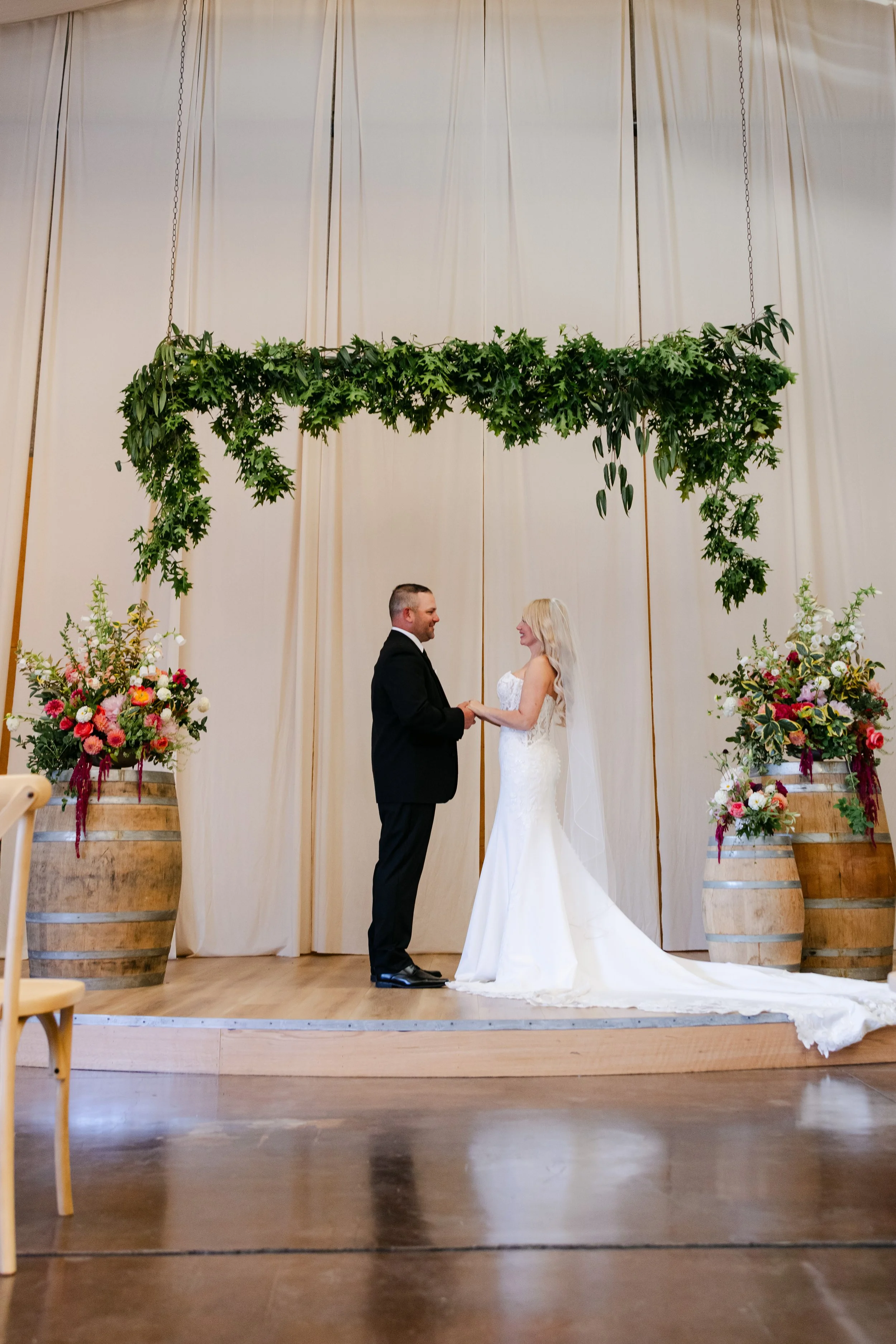 A wedding ceremony taking place indoors with a couple holding hands and looking at each other, dressed in wedding attire. There are tall floral arrangements and large wooden barrels on either side, with green foliage hanging above and a cream-colored