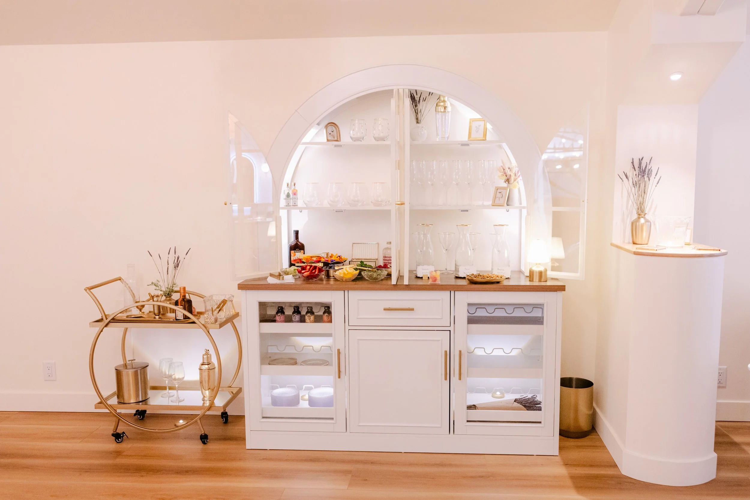 A white kitchen bar with glassware and decorative items on the shelves, a counter with food and drinks, and a gold bar cart with bottles and glasses. There is a small table with a plant in a white pot and a gold lamp, and a waste bin on the right sid