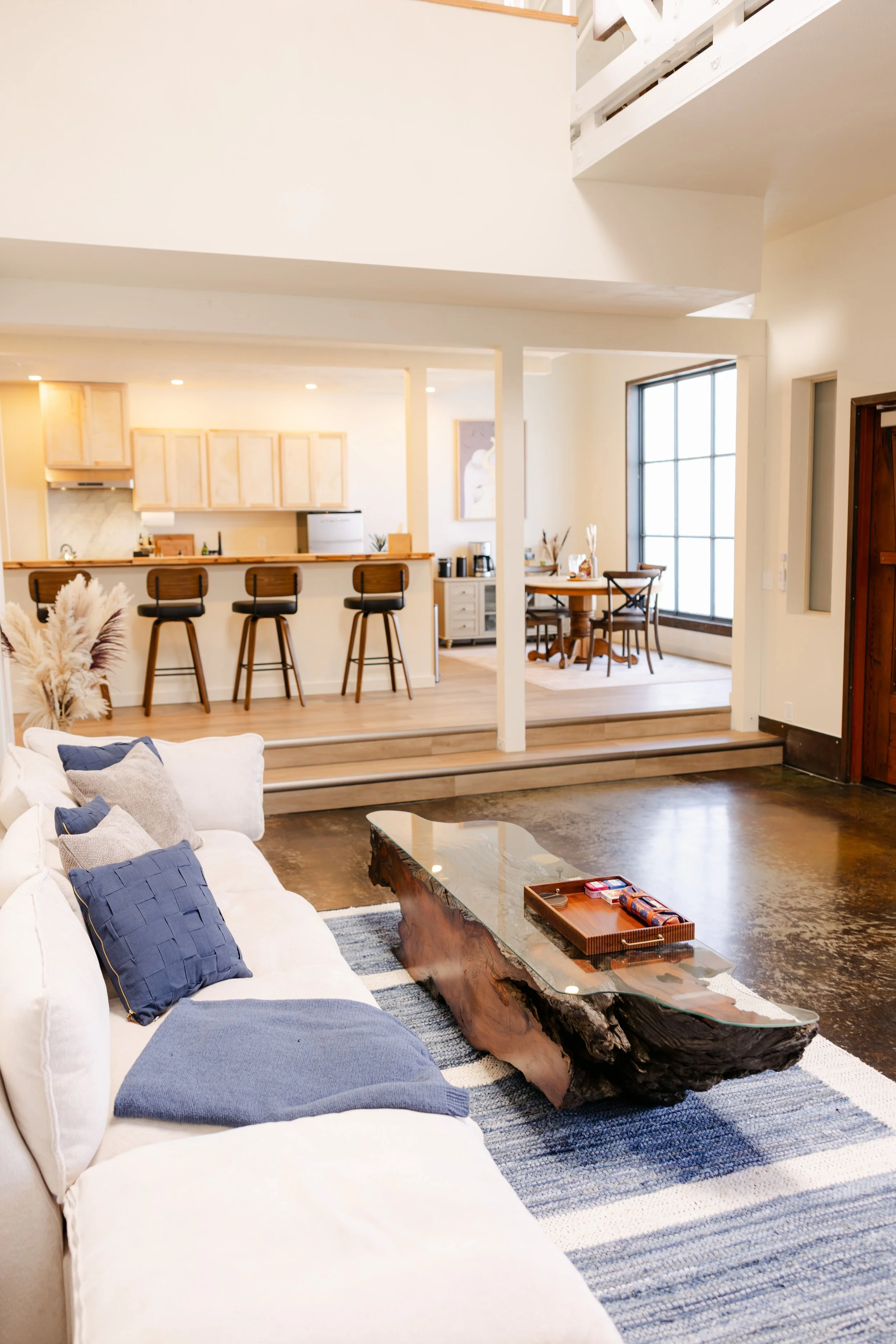 Living room with a white sofa, blue pillows, wooden coffee table, and a textured rug. Part of the kitchen and dining area with bar stools, a breakfast bar, and a large window with grid panes.