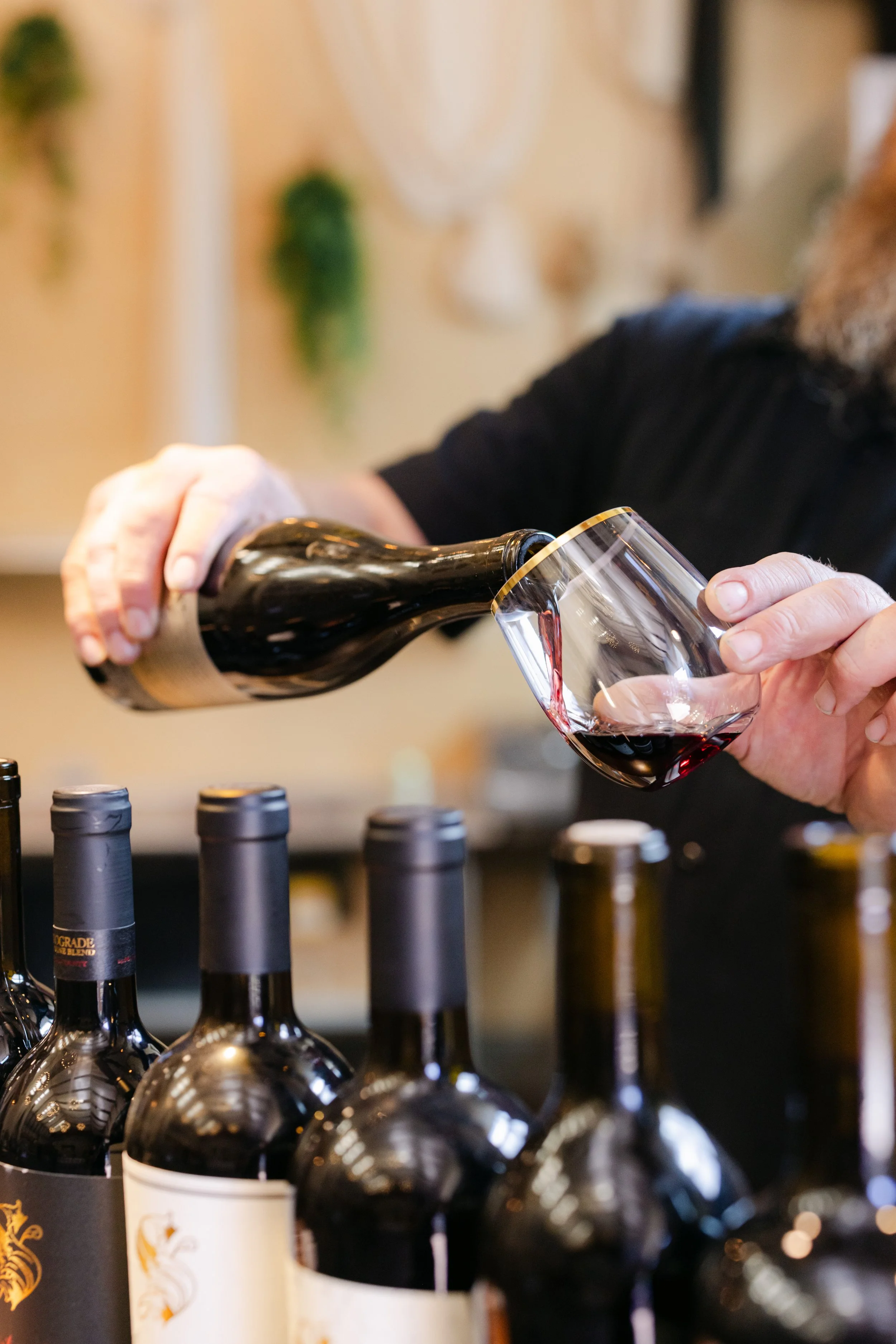 Person pouring red wine into a glass at a wine store or tasting event, with several wine bottles on display in the foreground.