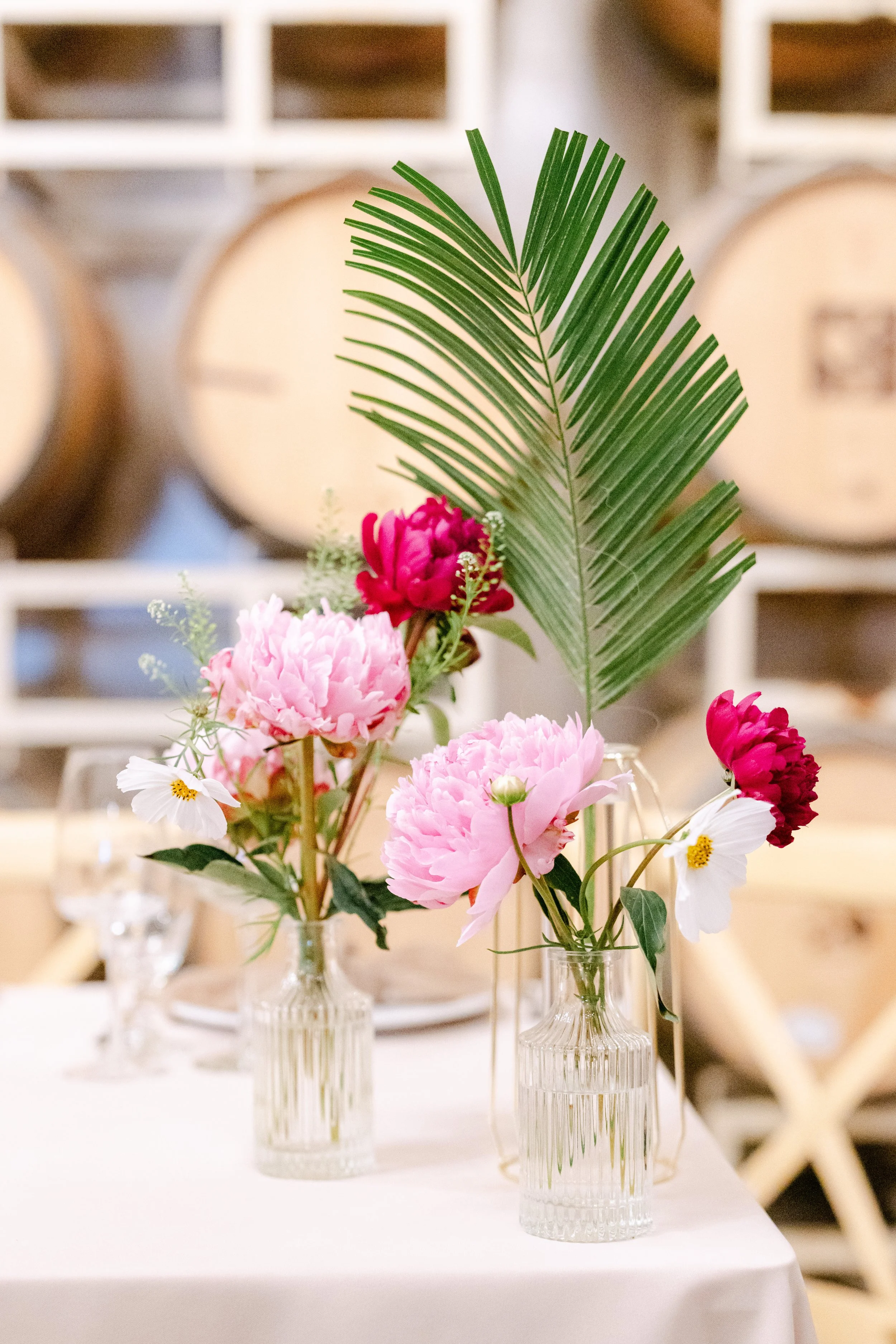 A floral centerpiece with pink and white flowers and large green palm leaves in three crystal vases on a white table, with a blurred background of barrels and shelving.