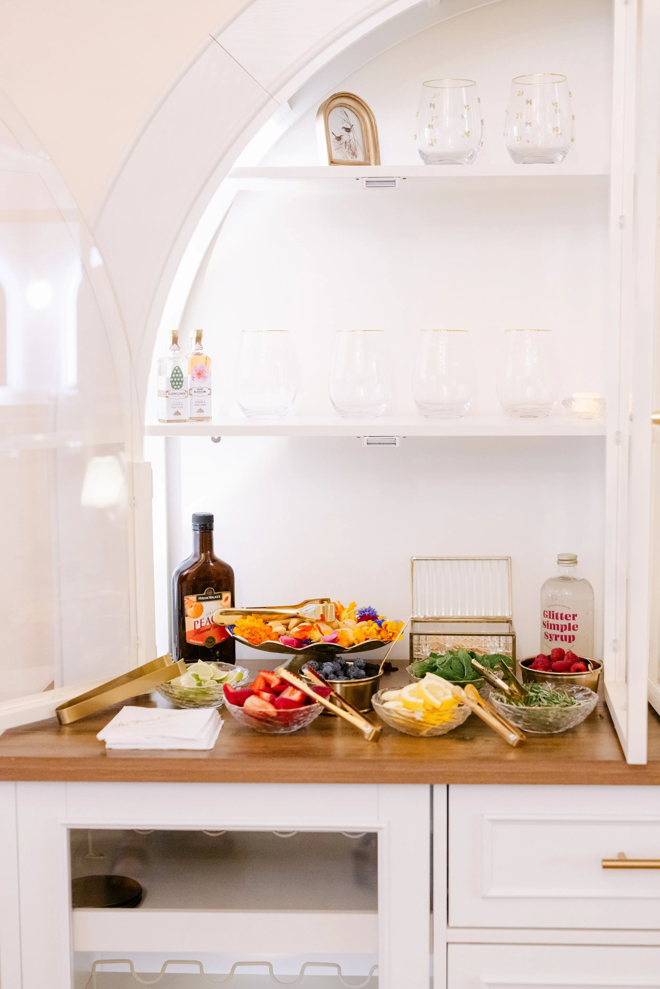 A white kitchen cabinet with open shelves and a counter filled with bowls of mixed fruits, including strawberries, blueberries, raspberries, and lemon slices, along with bottles of liquor and small items, under soft lighting.