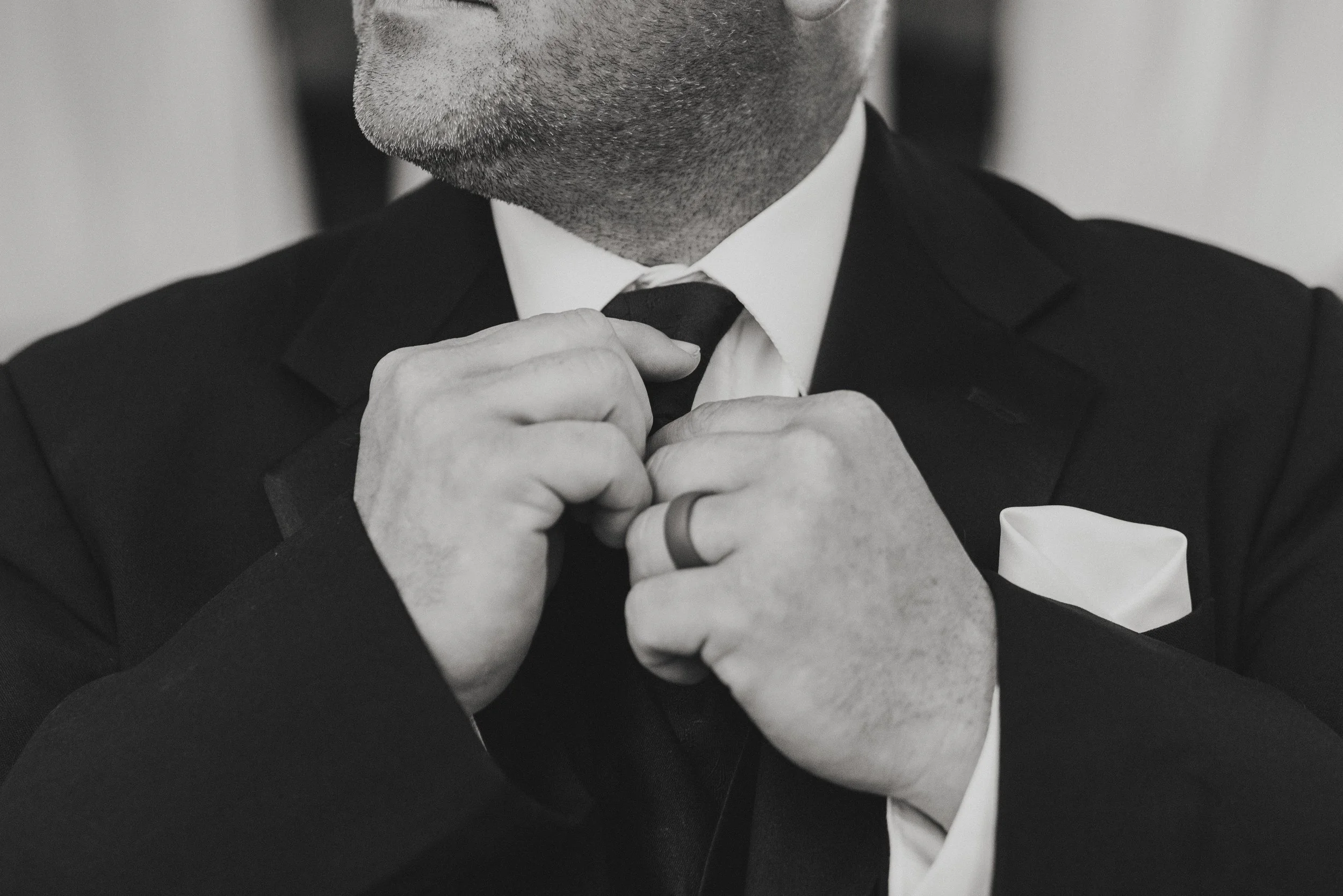 Close-up of a man adjusting his black tie, wearing a dark suit and a white shirt, with a blurred background.