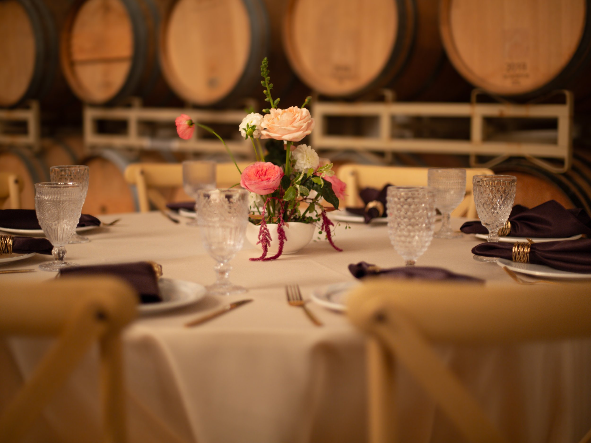 A dining table with a floral centerpiece, glasses, napkins, and silverware, set in a room with wine barrels in the background.