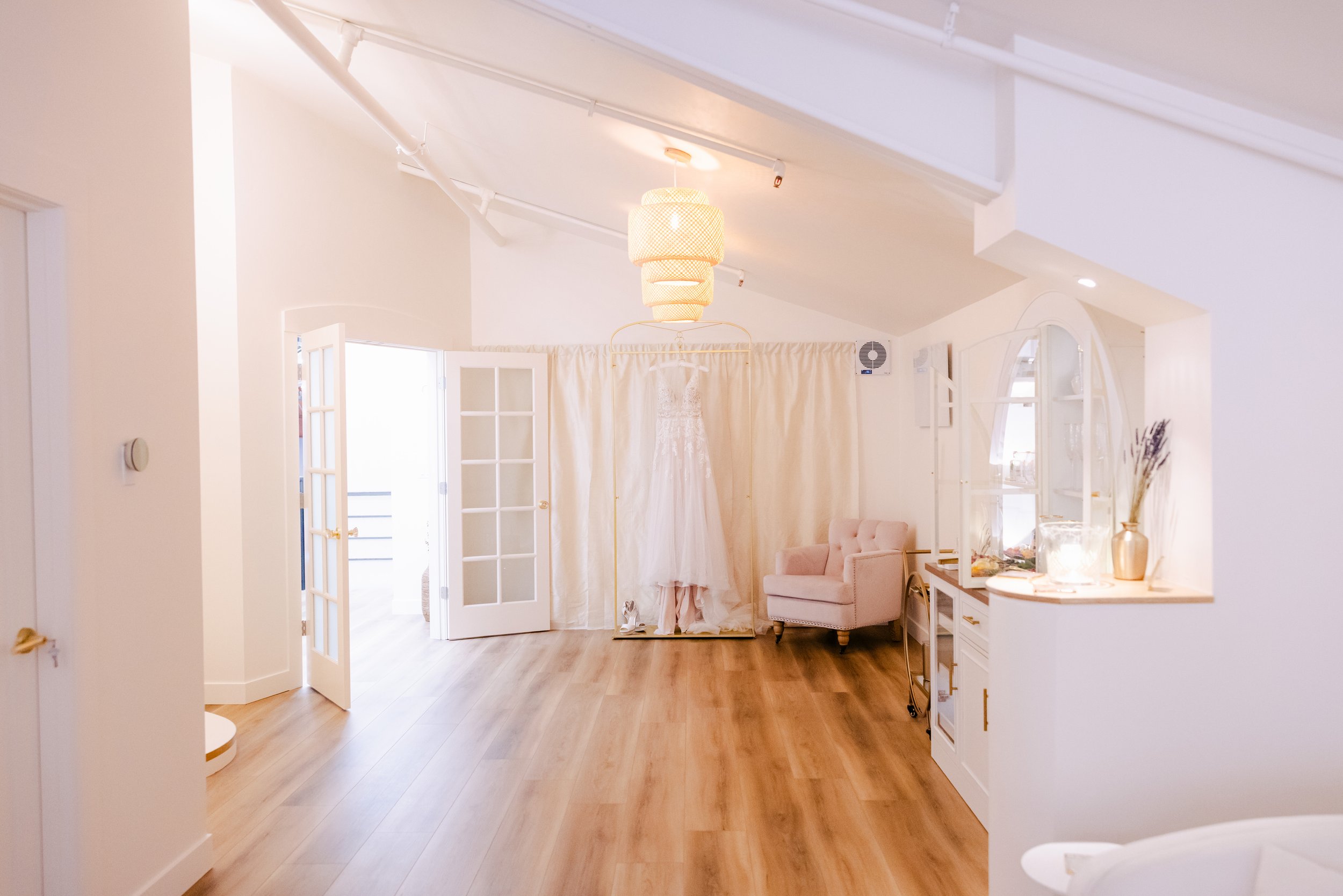 A bright, airy room with light-colored walls and wooden flooring. There is a white door with glass panes, slightly open, leading to an outside area. In the center, a hanging ceiling light with a woven lampshade illuminates the space. To the right, th