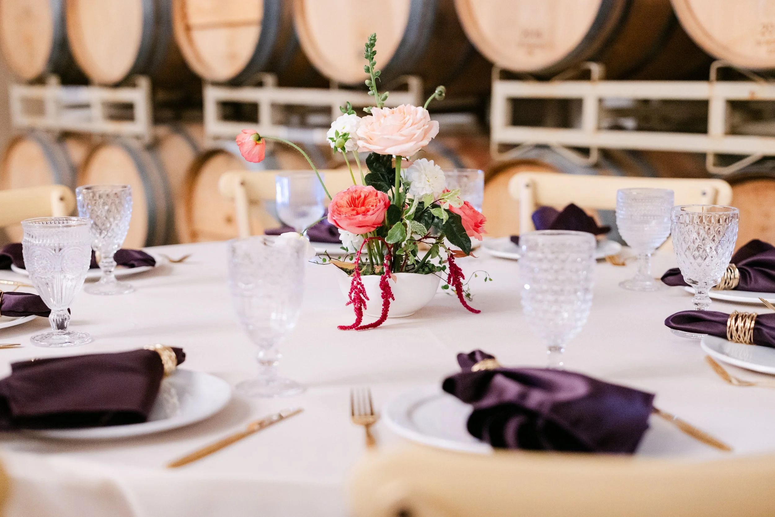 Table setting with a floral centerpiece in a winery or cellar, with wine barrels in the background.