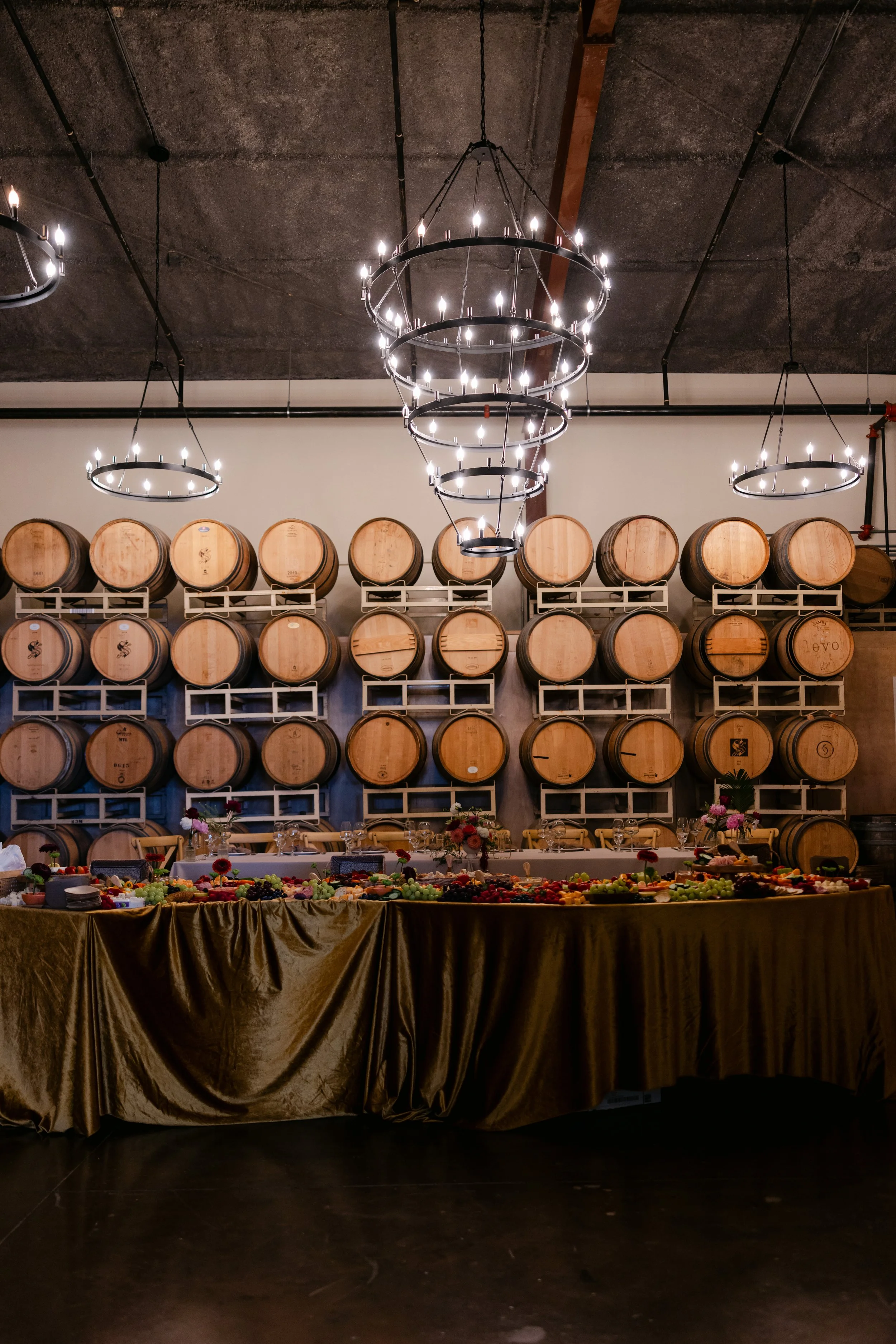 Wine barrels arranged on a rack on a wall behind a table with a fruit and cheese buffet, in a winery or event space with dark ceiling and modern chandeliers.