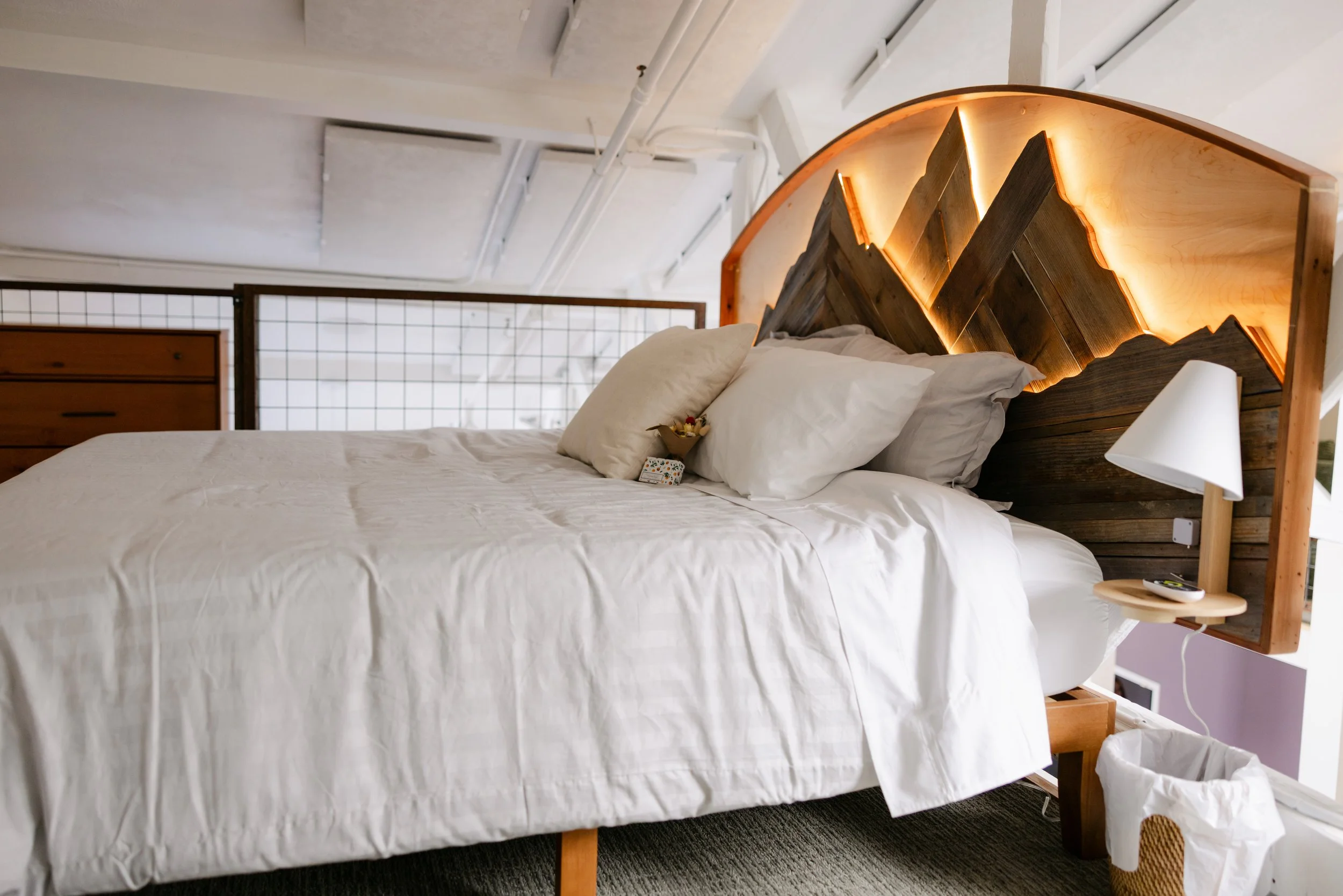 A neatly made bed with white bedding, two pillows, and a small flower arrangement on top, in a minimalist bedroom with modern decor, featuring a distinctive wooden headboard with backlit illumination, a bedside lamp, a wooden dresser, and a large win