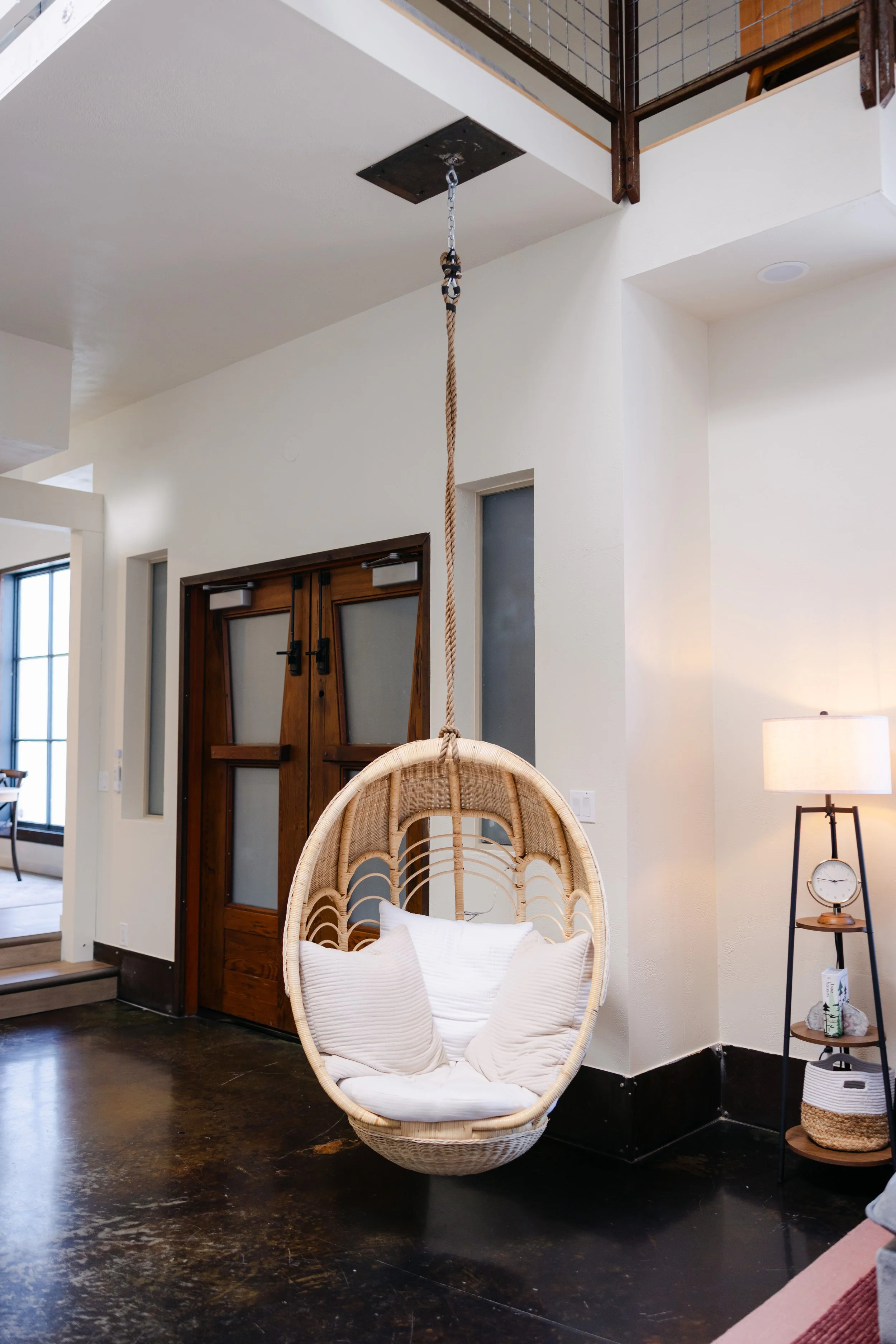 A cozy corner of a living space featuring a hanging circular wicker chair with white cushions, situated on a dark wooden floor. Behind the chair, there's a wooden door with frosted glass panels, and to the right, a side table with a clock and decorat