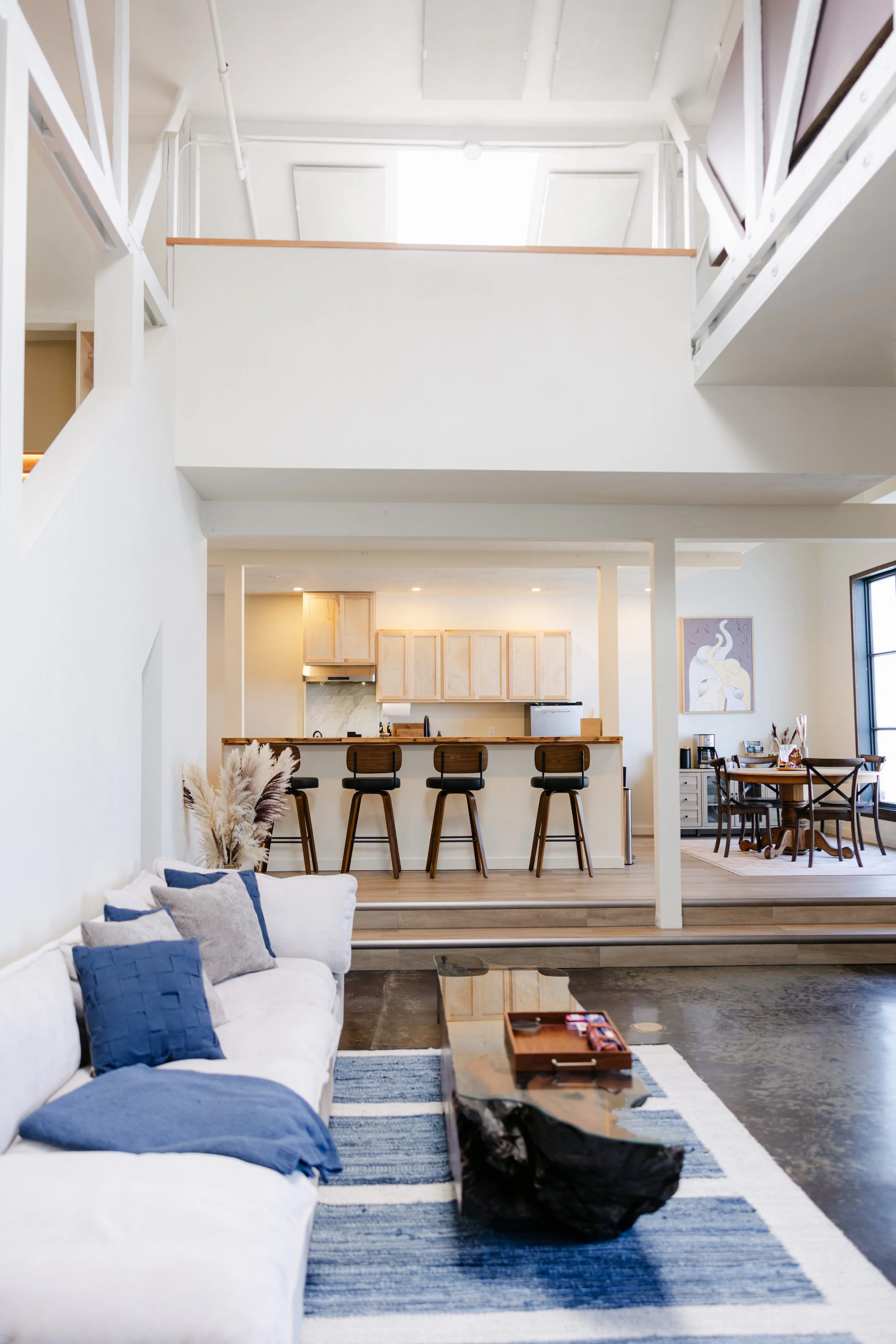 Open-concept living and kitchen area with white sectional couch and blue pillows, wooden coffee table, blue and white striped rug, bar stools at kitchen counter, wooden cabinets, and artwork on wall.