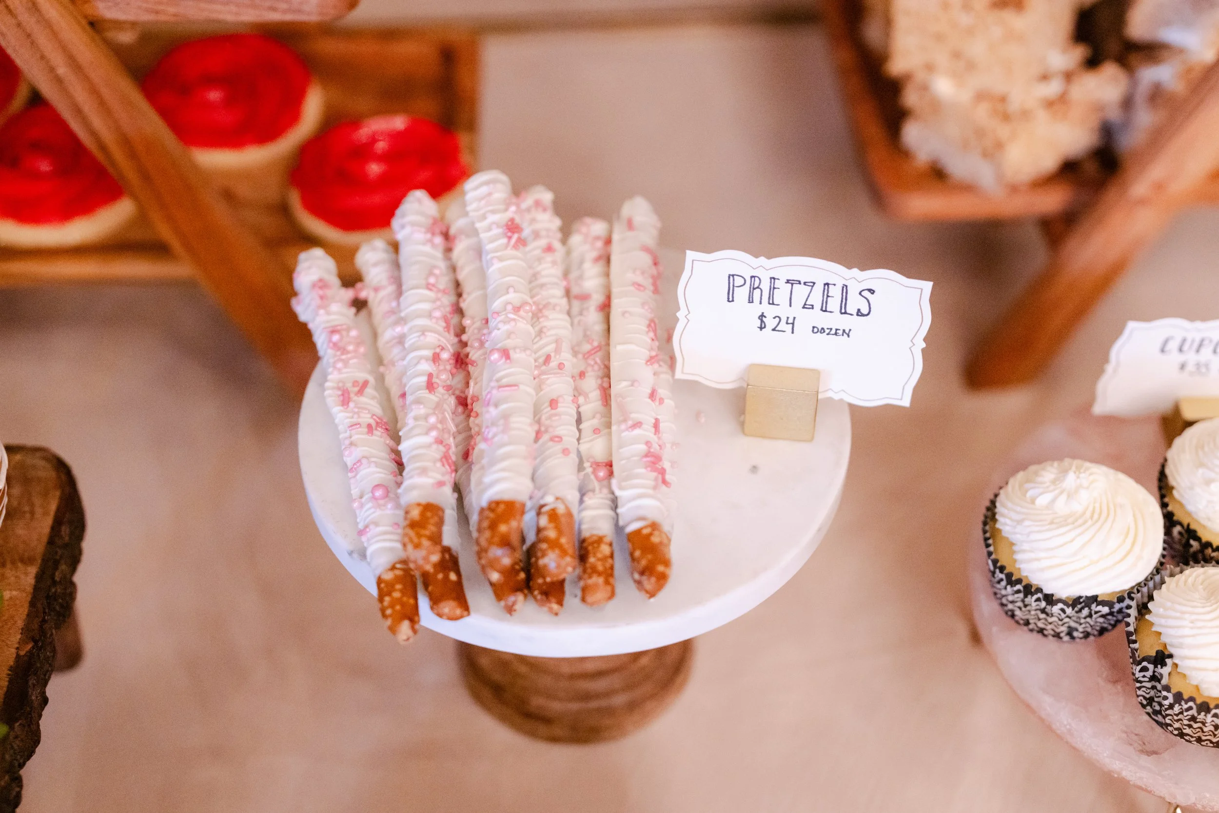 Plate of pretzels decorated with pink and white icing and sprinkles with a sign showing the price of $24 per dozen.