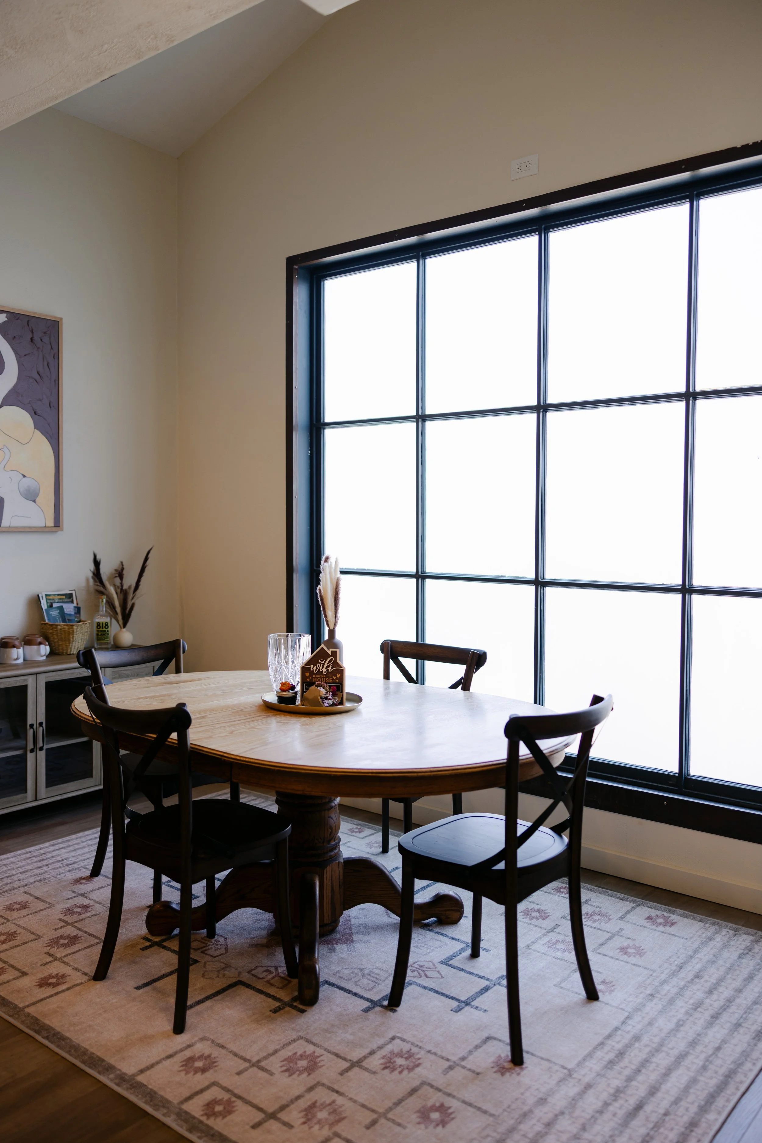 A cozy dining room with a round wooden table and six chairs, a large window with black frames letting in natural light, a patterned area rug, a sideboard with decorative items, and a hanging painting on the wall.