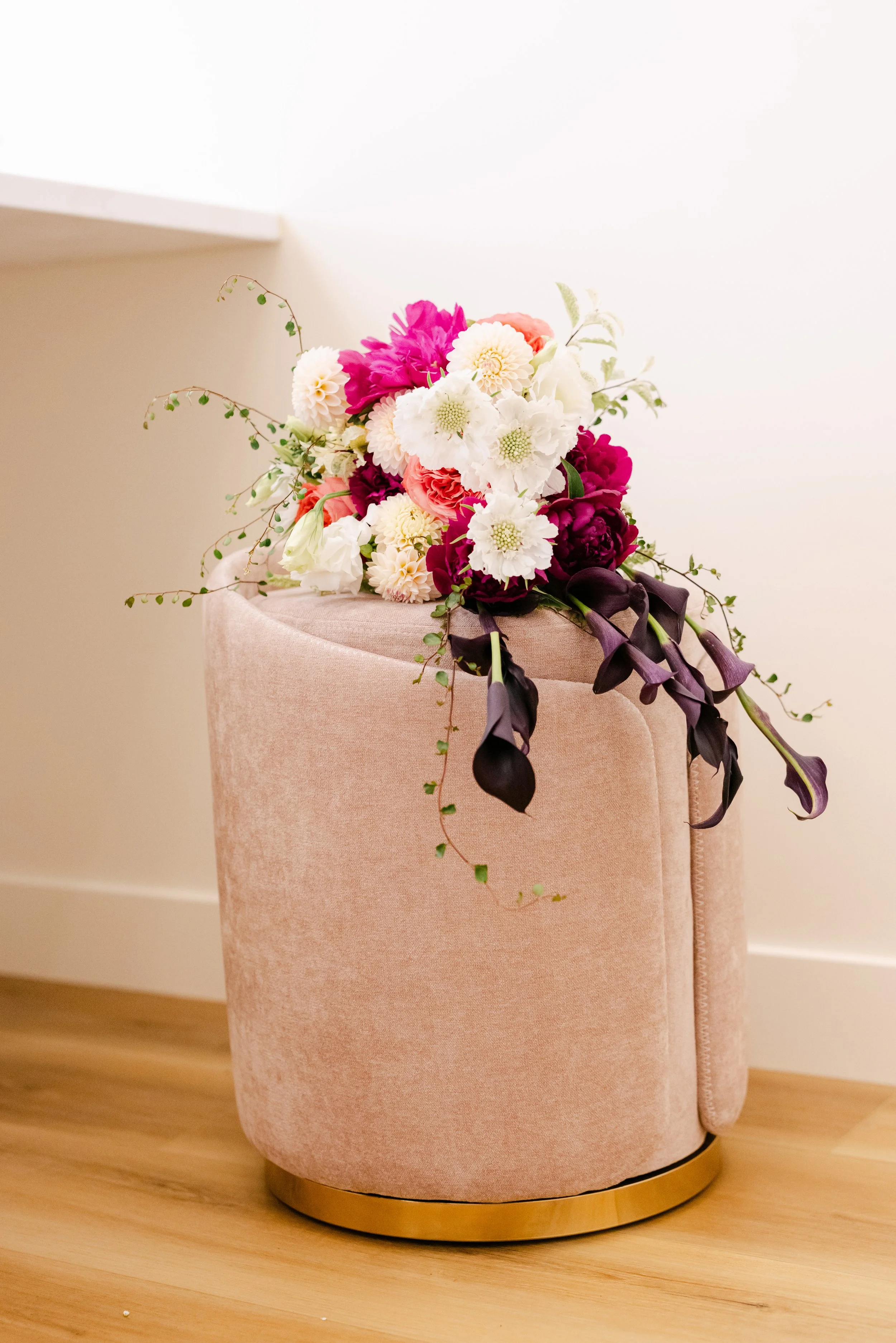 A bouquet of pink, white, and dark purple flowers resting on a pink upholstered chair with a gold base, placed on a wooden floor against a plain wall.