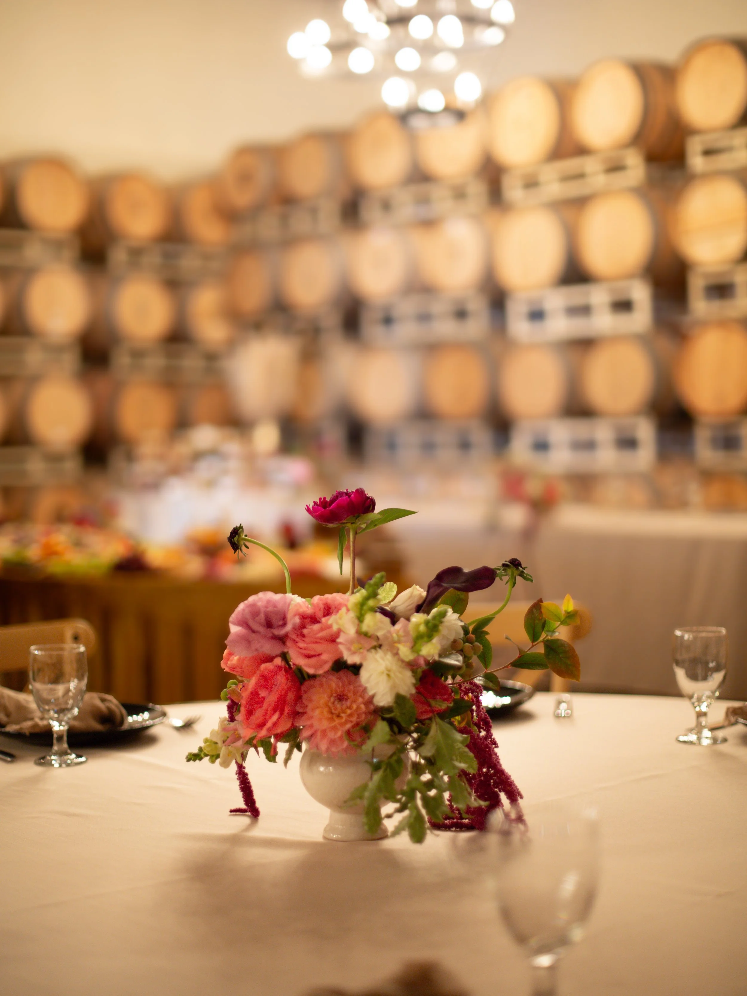 A floral centerpiece featuring pink, red, and white flowers on a banquet table, with a blurred background of wine barrels and warm lighting.