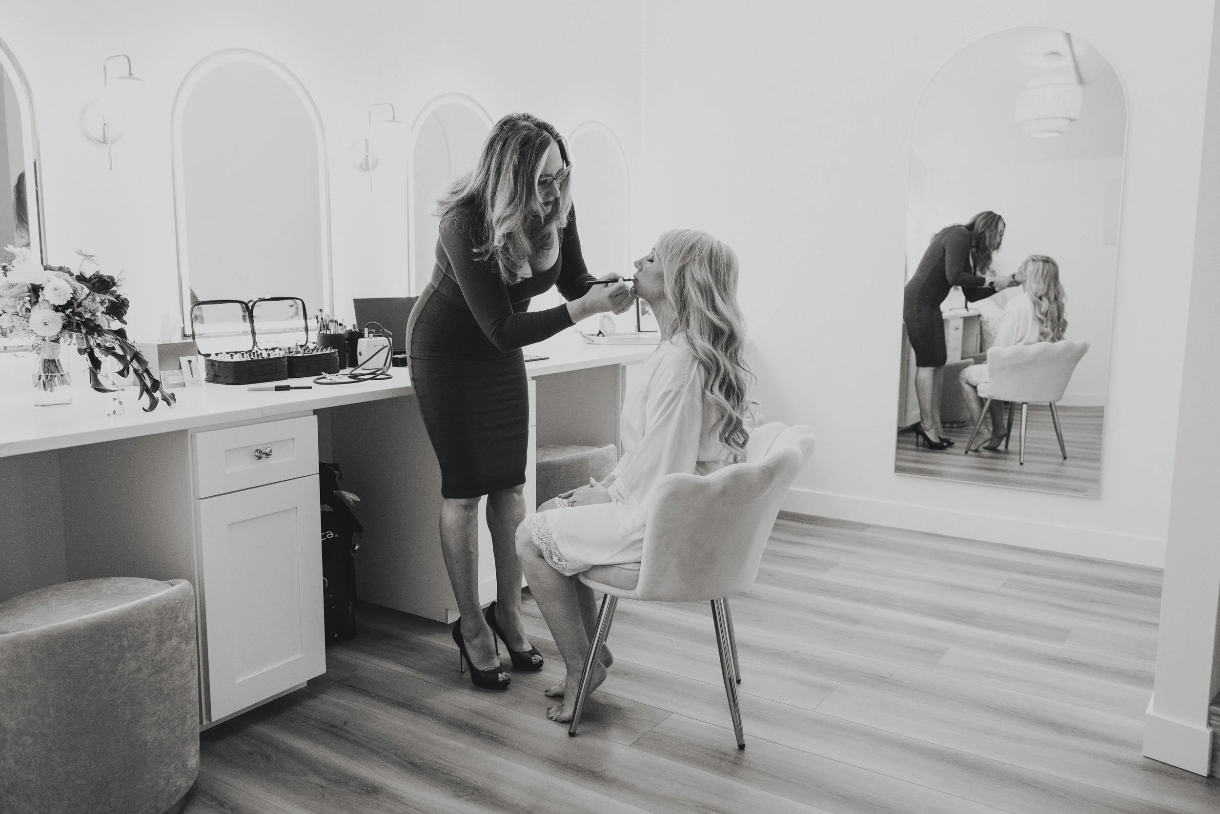 Makeup artist applying makeup to a woman seated in a chair in a bright, modern room. Reflection in a mirror shows the scene.
