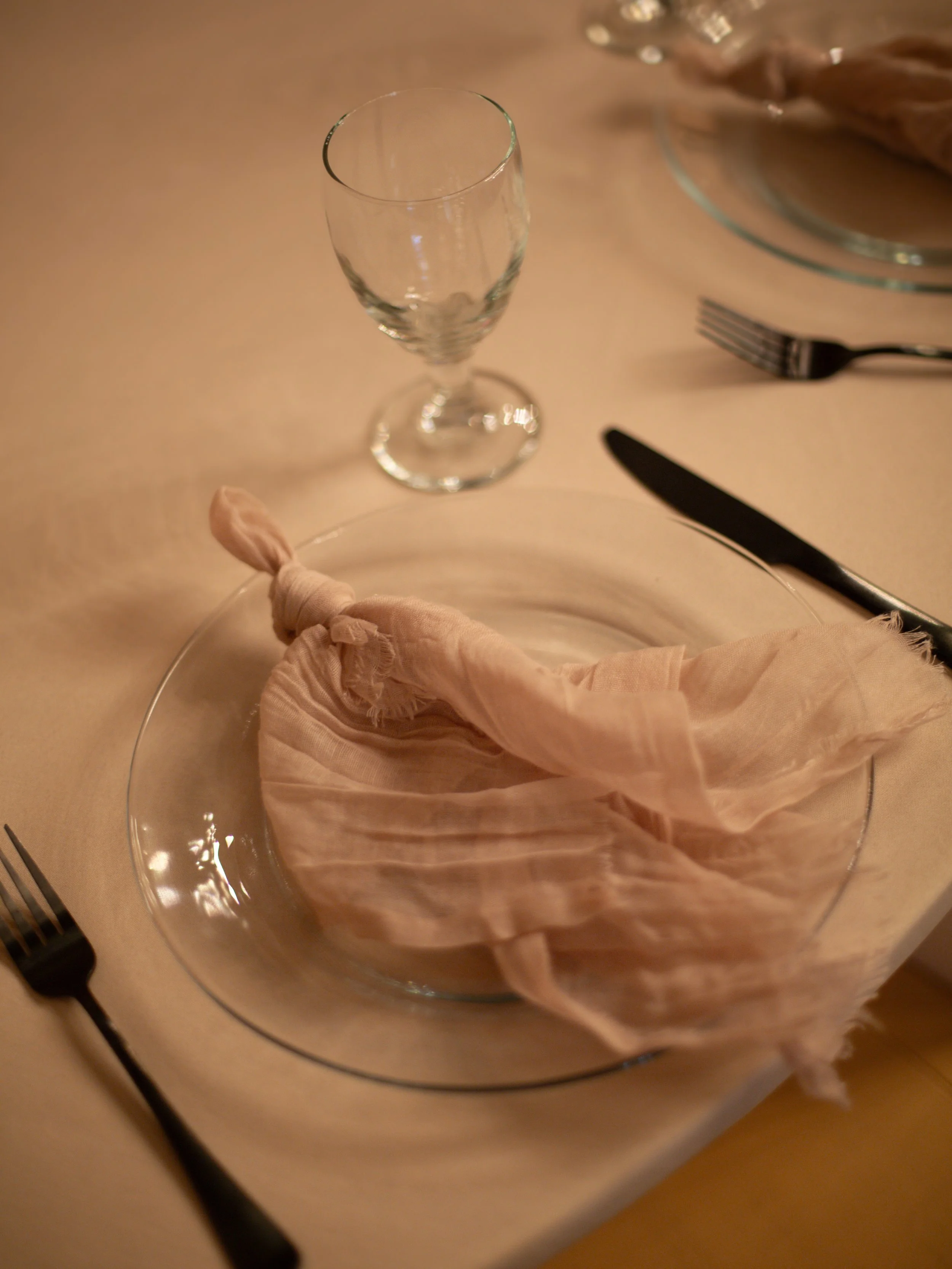 Empty wine glass on a table next to a plate with a tied cloth napkin, a fork, and a knife. A bowl with some food is visible in the background.