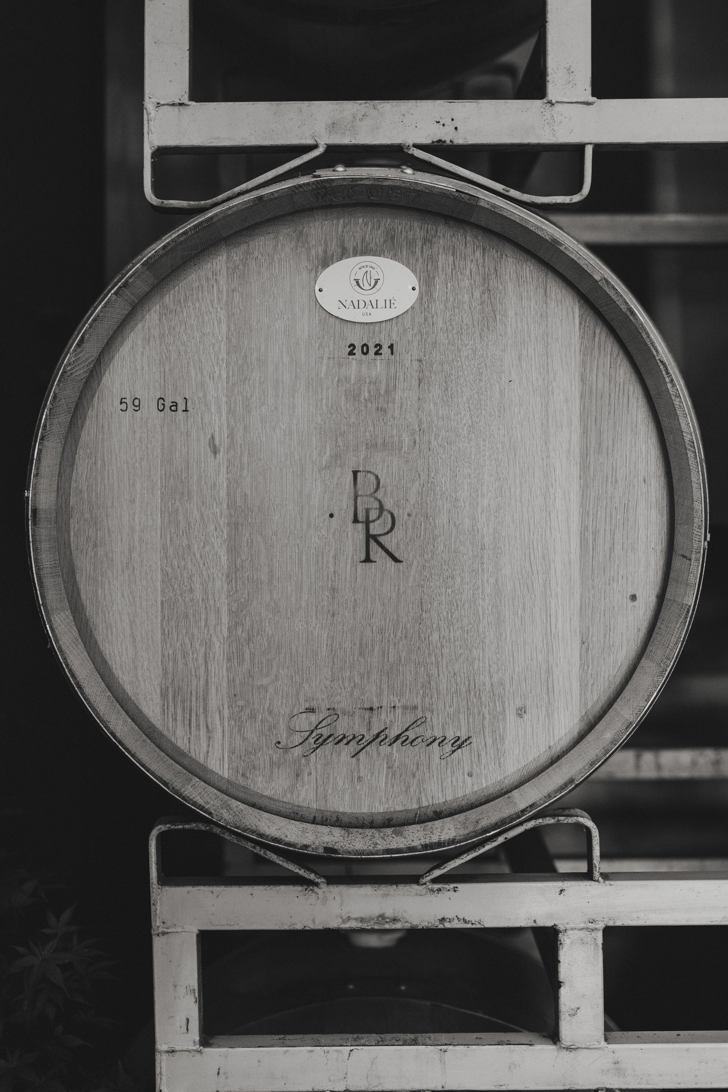 A wooden wine barrel with a metal band, placed on a white wine rack. The barrel has labels that say 'Nadalié,' '2021,' and '59 Gal,' with the word 'Symphony' written at the bottom.