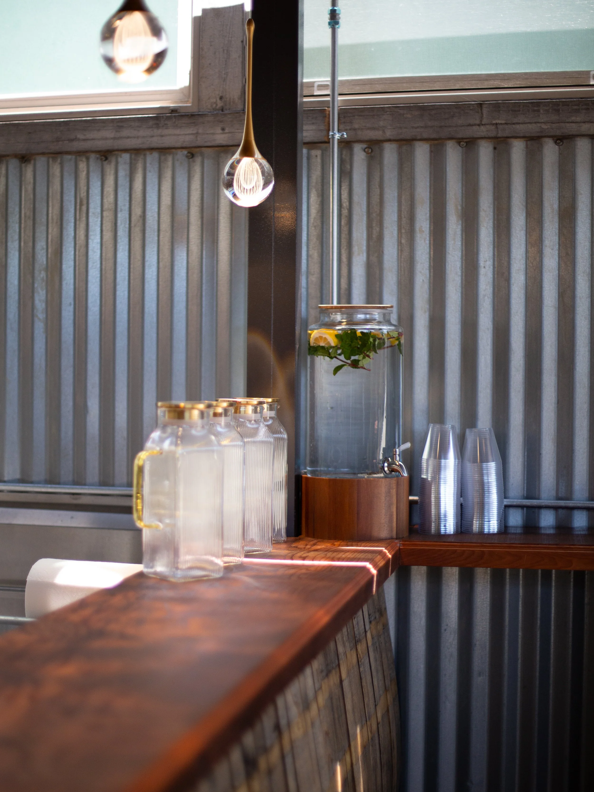 A wooden bar counter with glass beverage dispensers and cups, and a large jar of infused water with lemon and mint, in a rustic setting with metal wall paneling and a window.