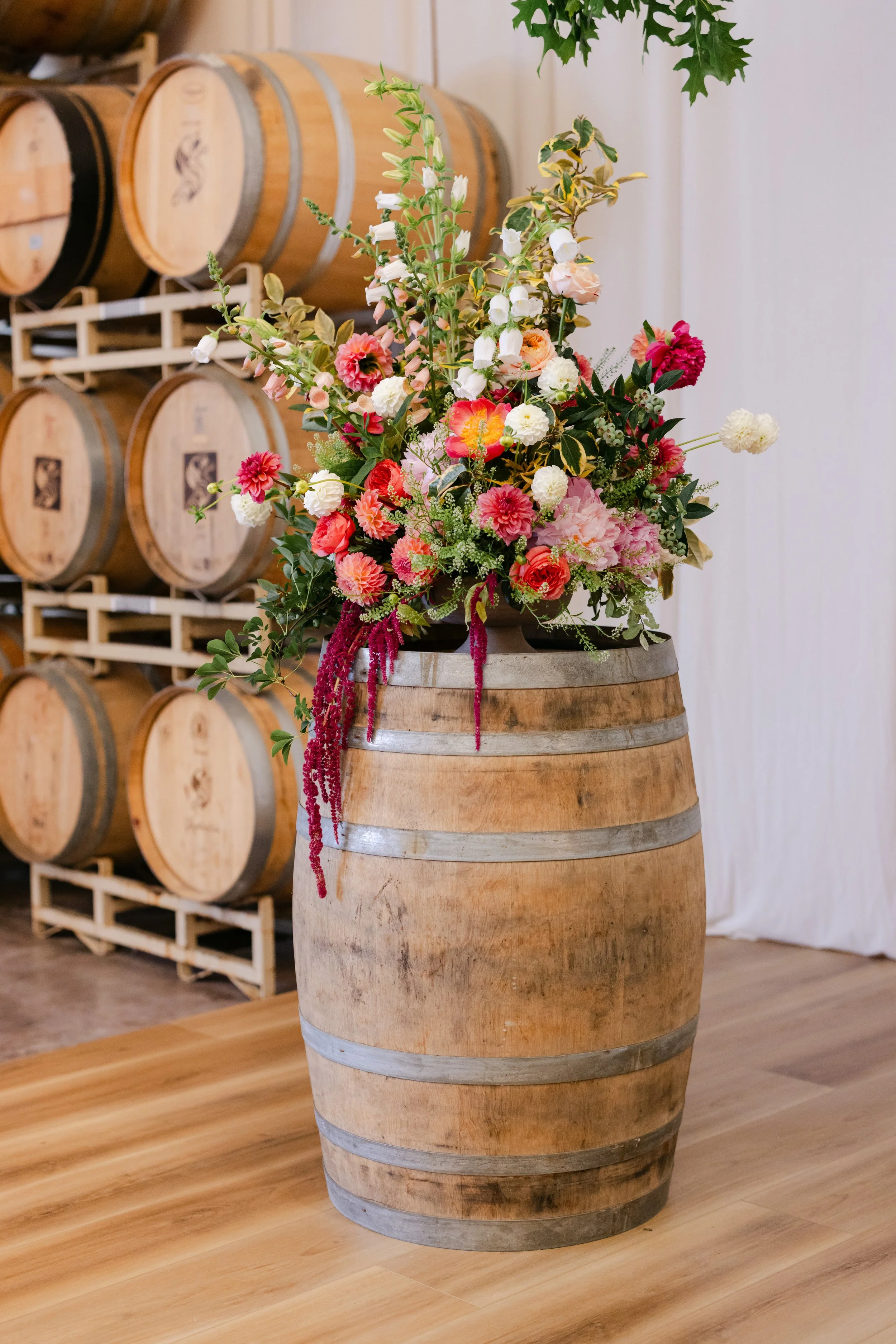 A barrel with a bouquet of colorful flowers on top, set in a wine cellar with more barrels in the background.