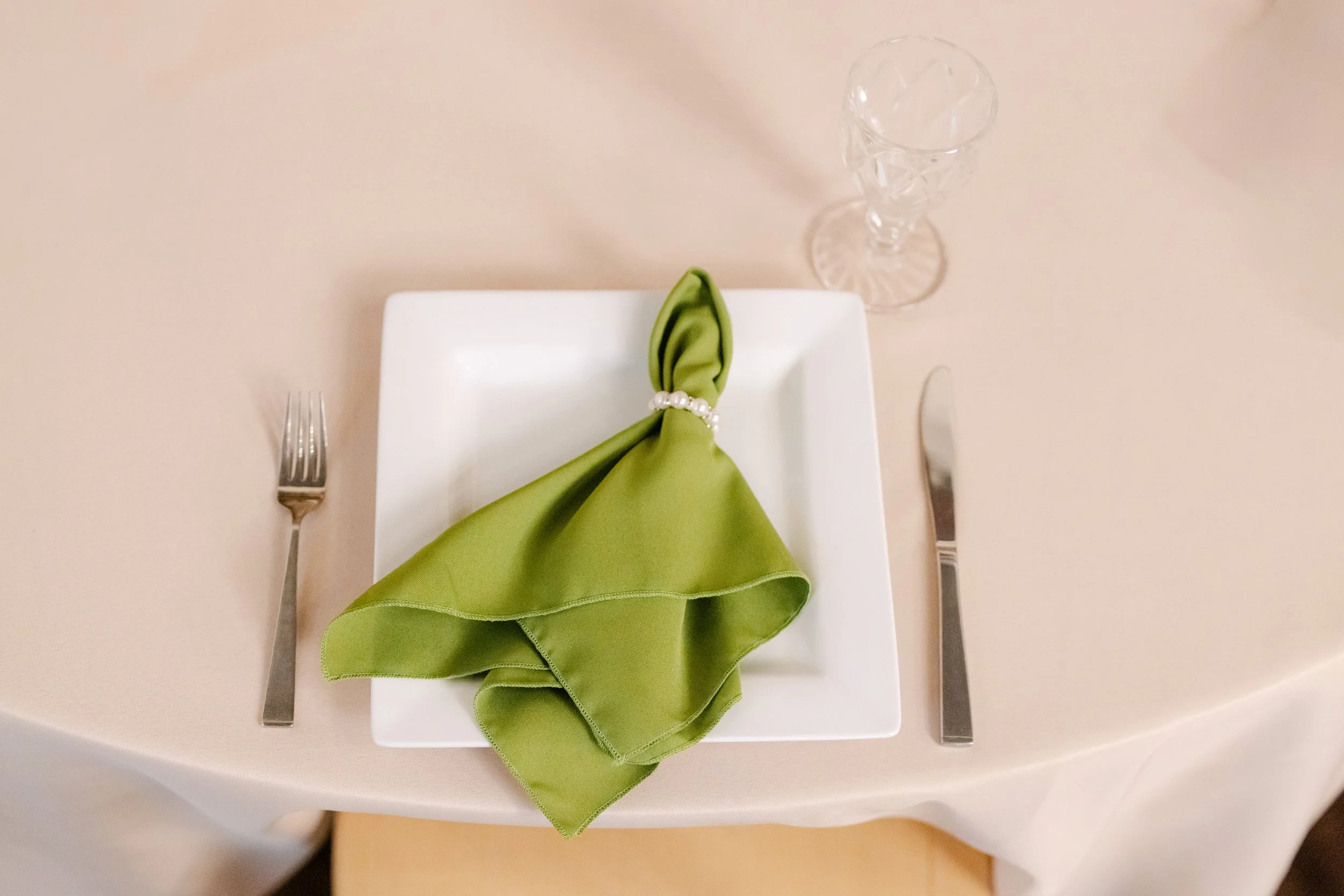 Place setting on a dining table with a white square plate, a green cloth napkin folded and secured with a pearl napkin ring, a fork on the left, a knife on the right, and a tall, empty crystal wine glass at the top right.