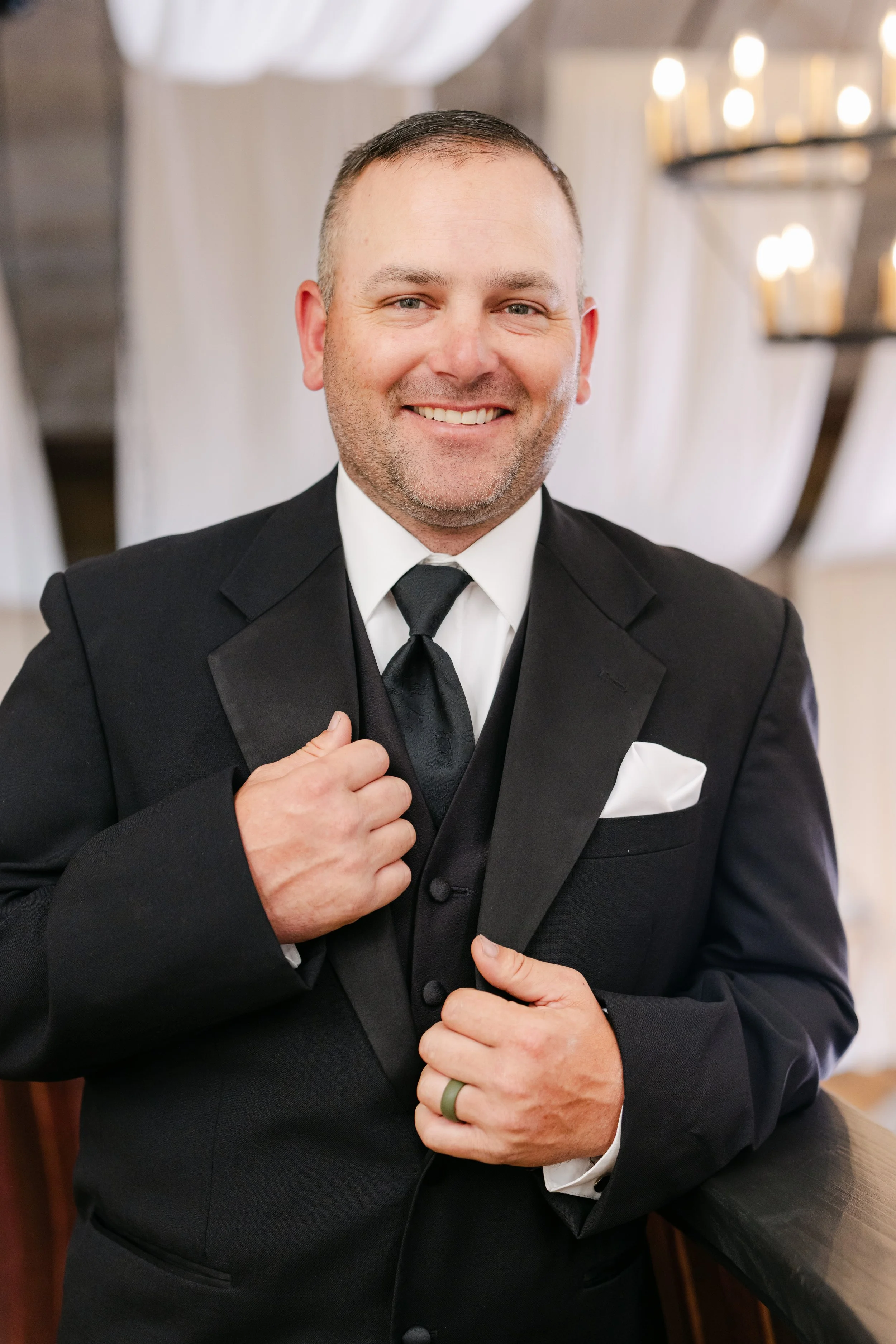 A smiling man dressed in a black tuxedo with a black tie and a white pocket square, standing indoors in a formal setting.