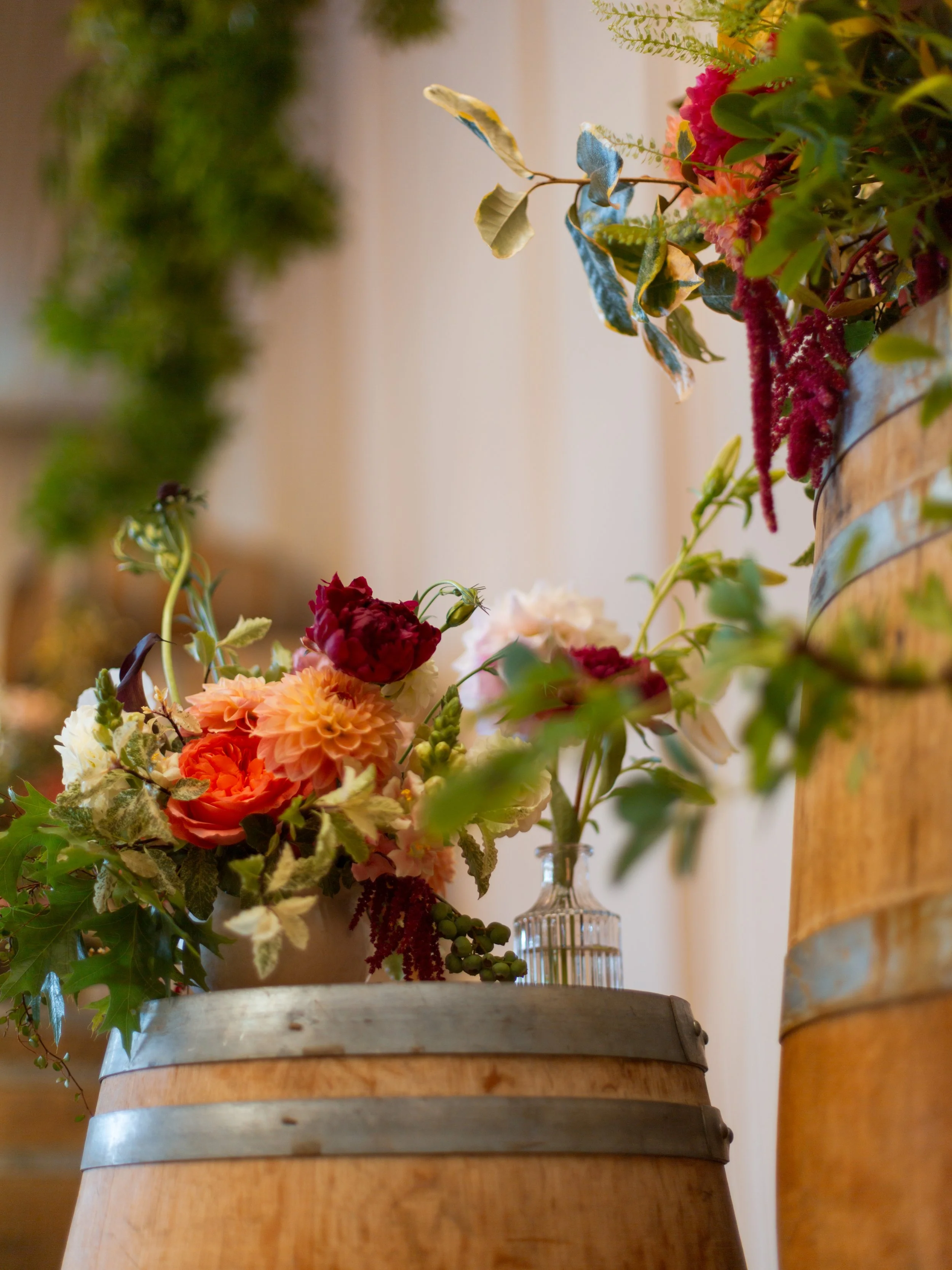 Vase holding colorful flowers on a table, with a wooden barrel and greenery in the background.