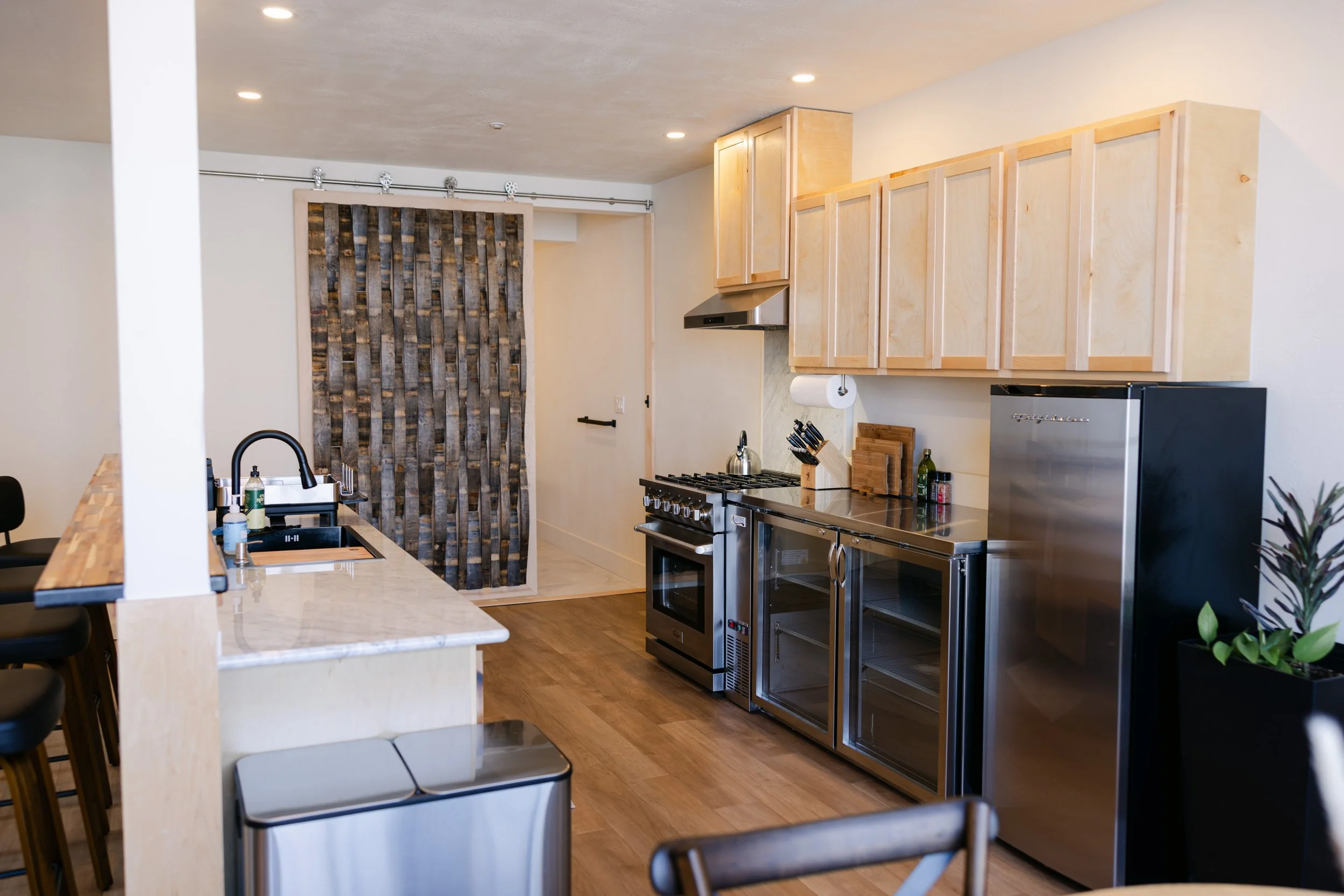 Modern kitchen with beige cabinets, stainless steel appliances, a black stove, a wine fridge, and a marble-topped island with black barstools.