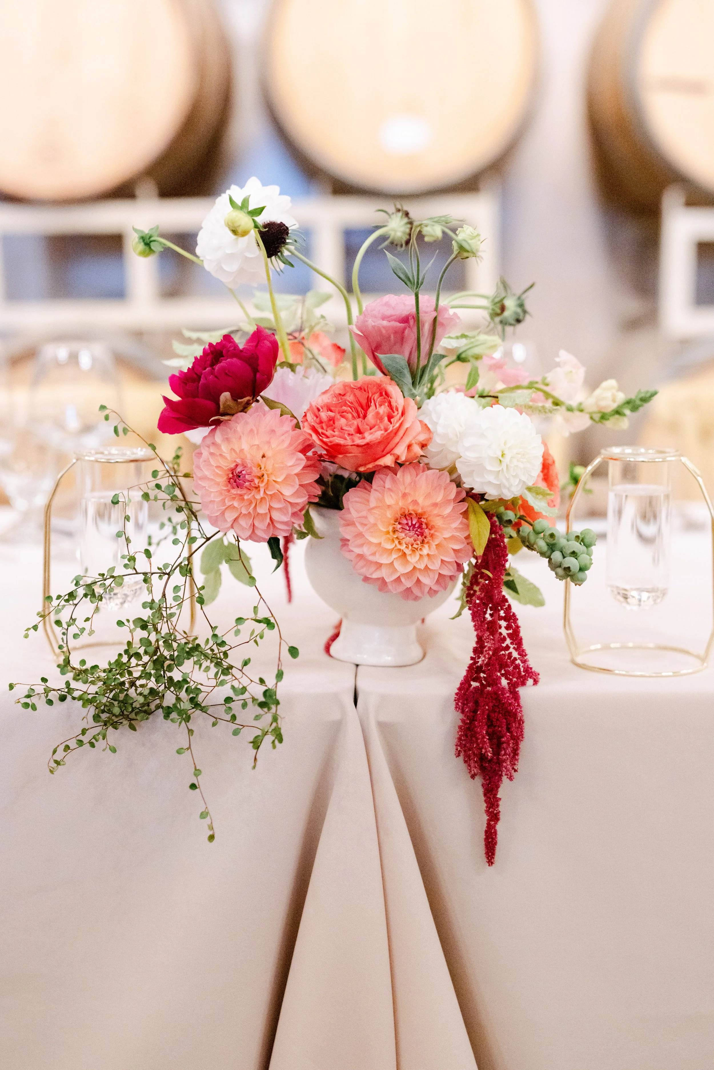 A floral centerpiece with pink, white, and red flowers, accented with green foliage, placed on a white table with glassware and surrounded by a beige tablecloth and beige walls.