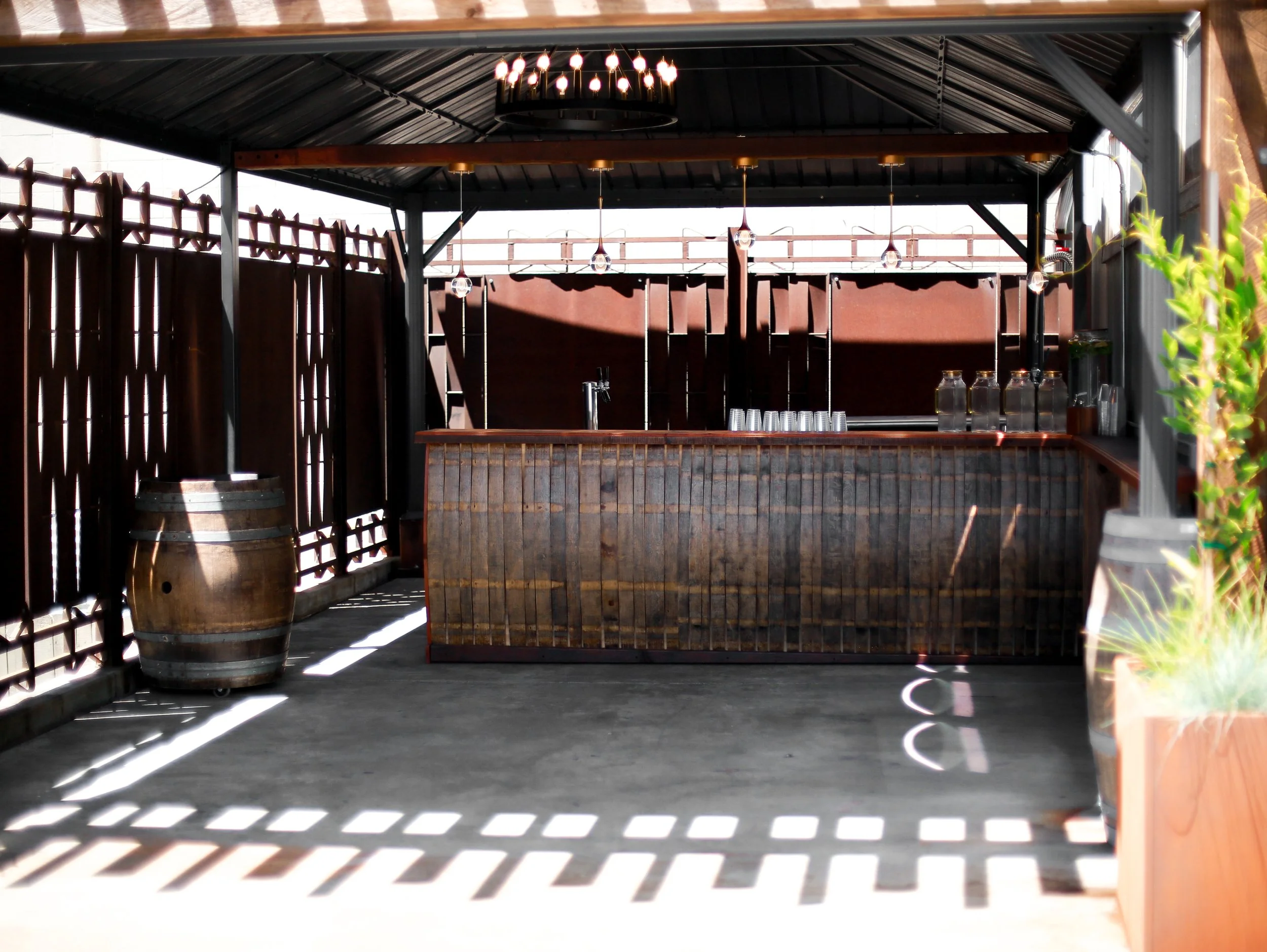 Outdoor bar area with wooden bar counter, hanging pendant lights, a circular chandelier, an empty barrel, and a wooden fence with sunlight casting shadows.