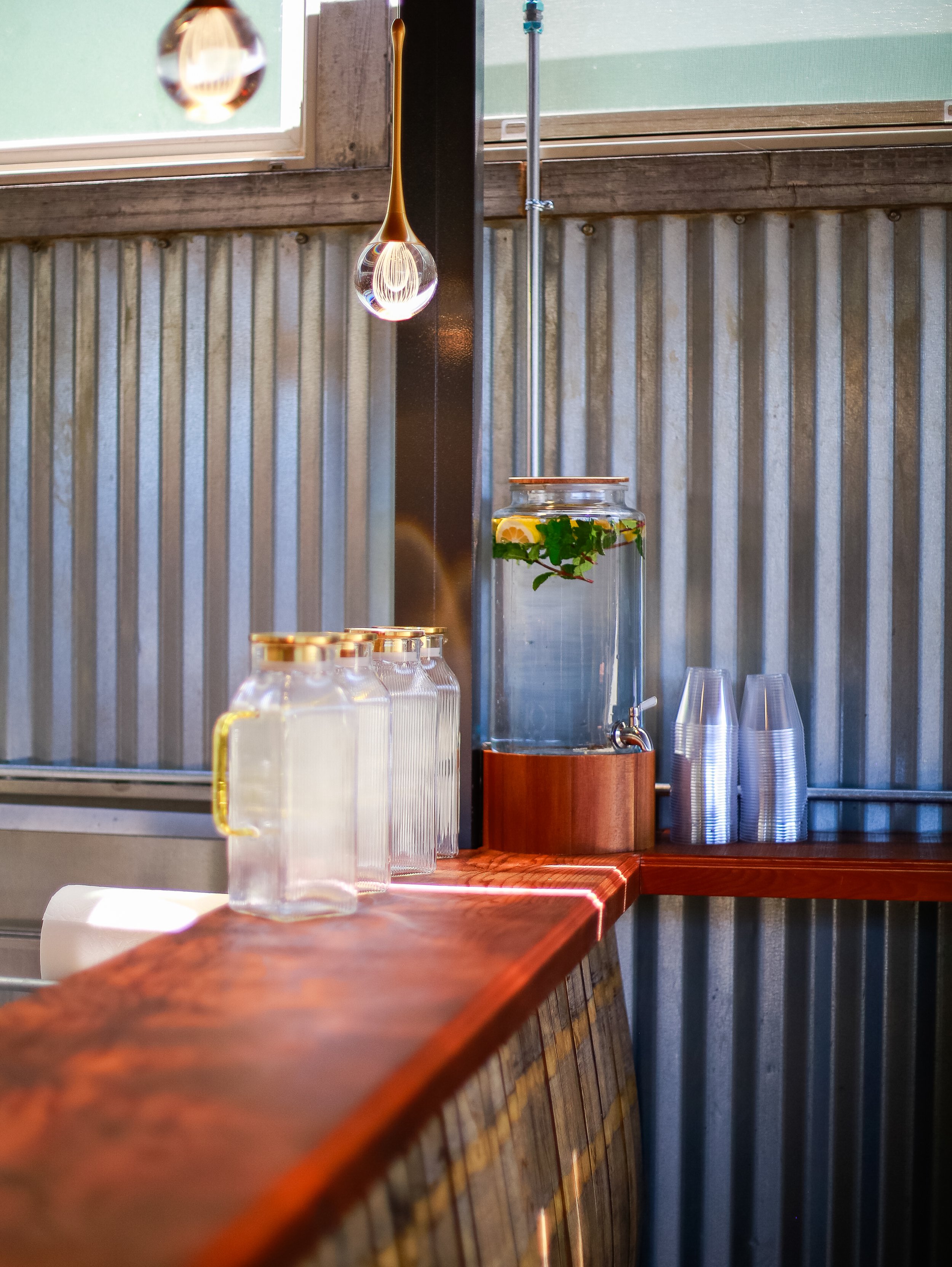 A wooden counter with a glass jar filled with water, fruit slices, and herbs, a water dispenser, and clear plastic cups stacked on the side. A window above with a water droplet hanging on the frame.