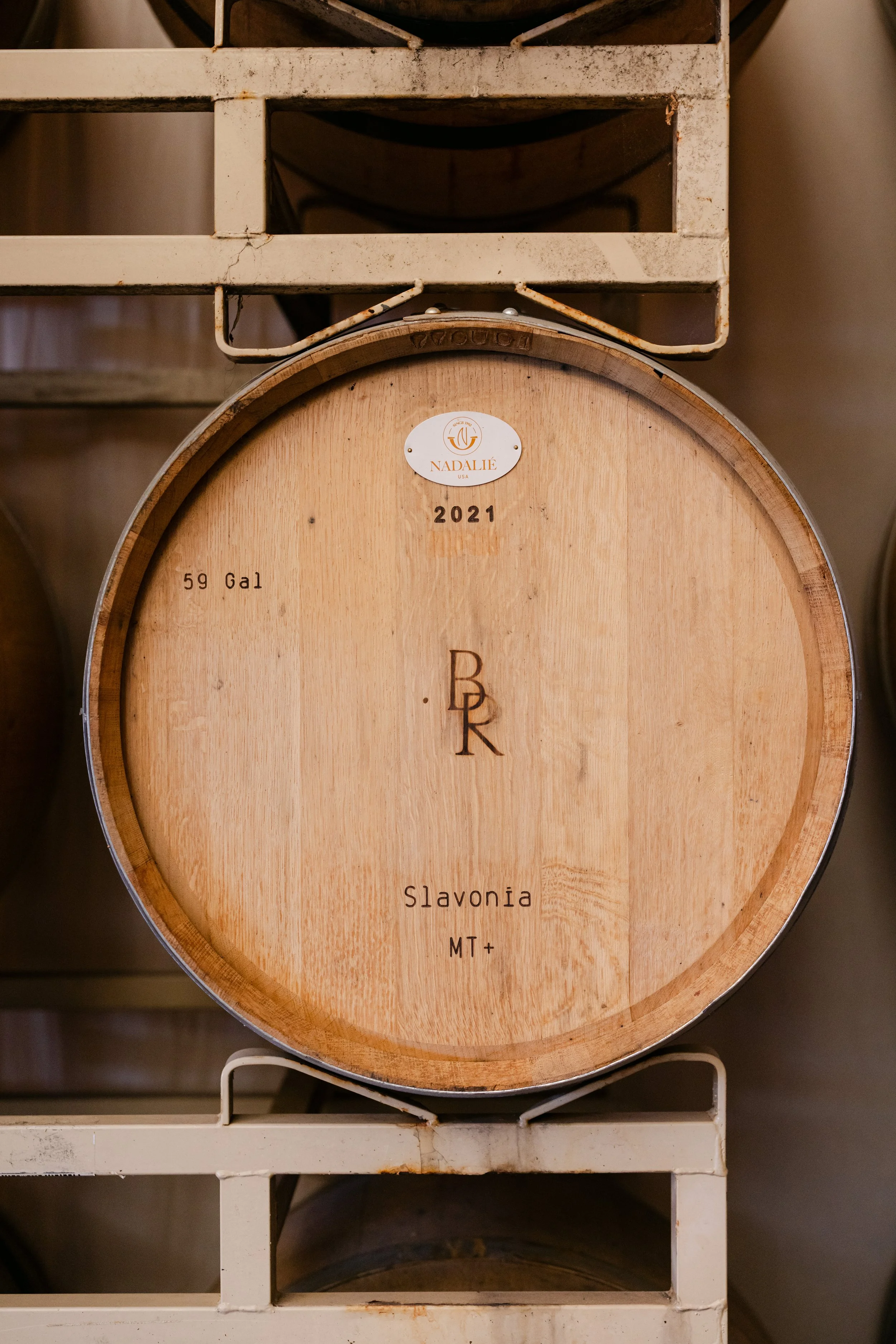 A wooden barrel with a metal band around it, labeled with the brand 'Navalie' and the year 2021. The barrel features the text 'Slavonia MT+' and the volume '59 Gal'. The barrel is placed on a white metal rack, with some rust visible on the rack.