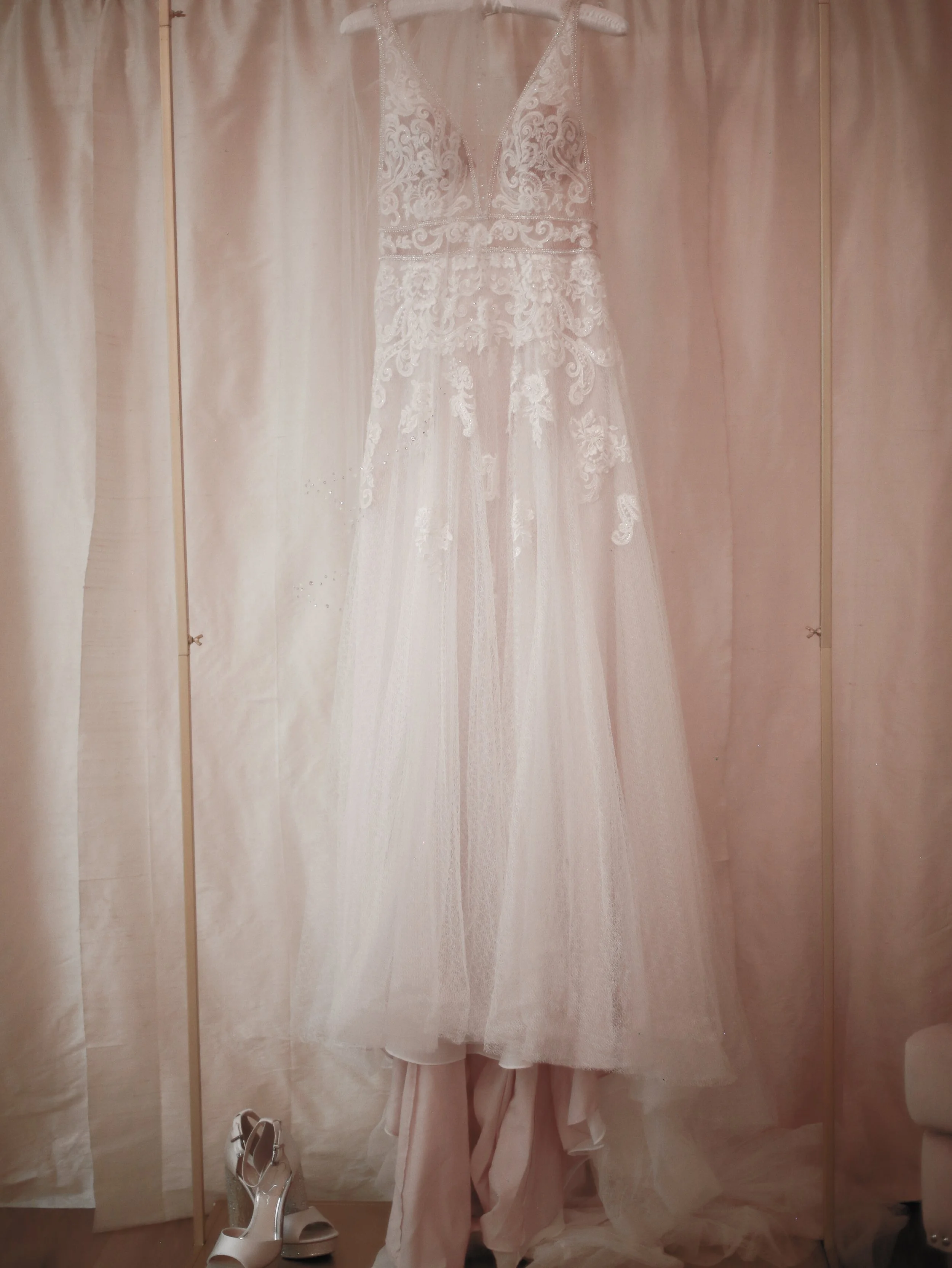 A wedding dress hanging on a hanger in front of a pink curtain, with a pair of white high-heeled shoes on the floor below.