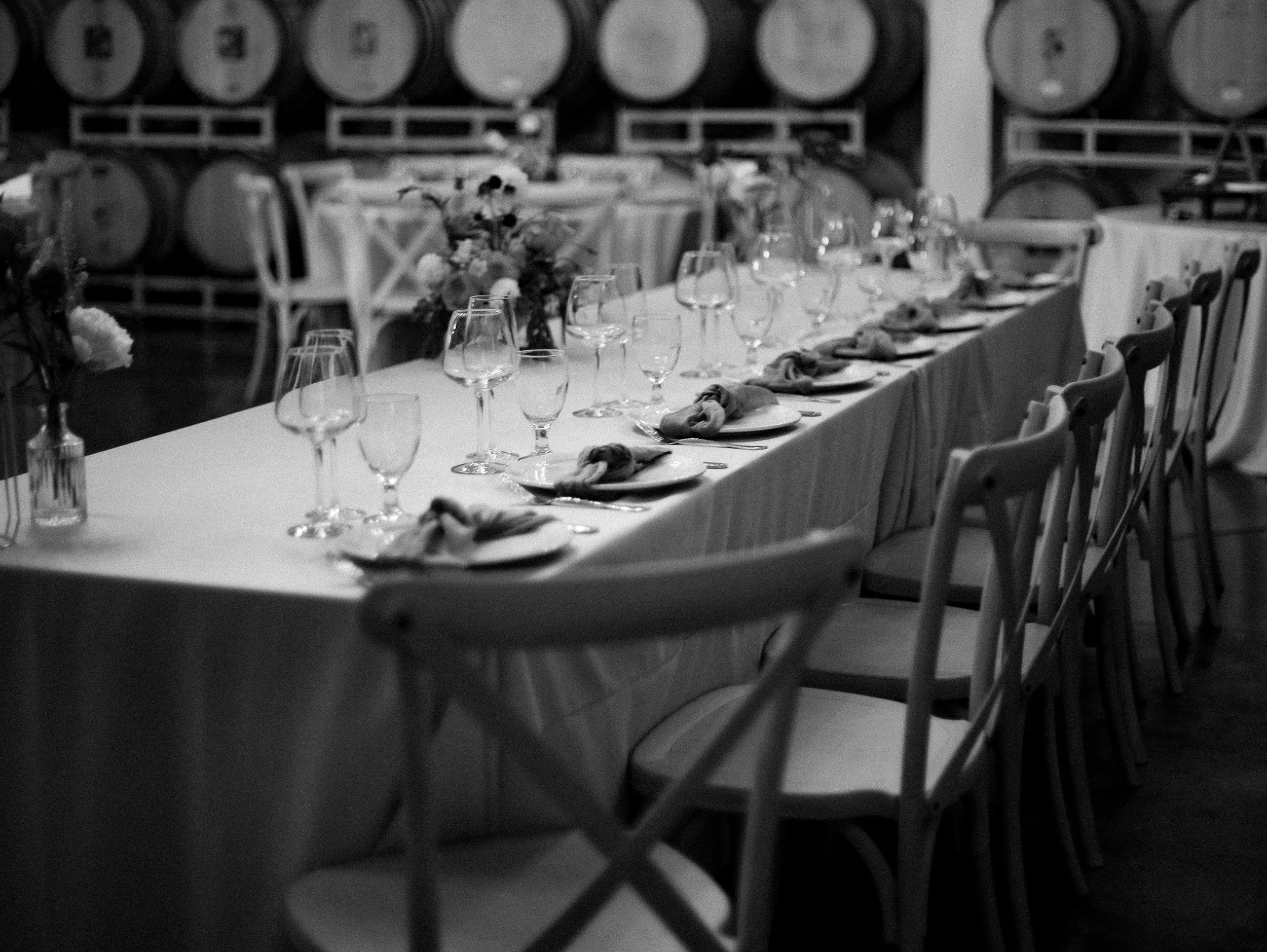 Black and white photo of a long dining table set with wine glasses, plates, and napkins, with wine barrels in the background.