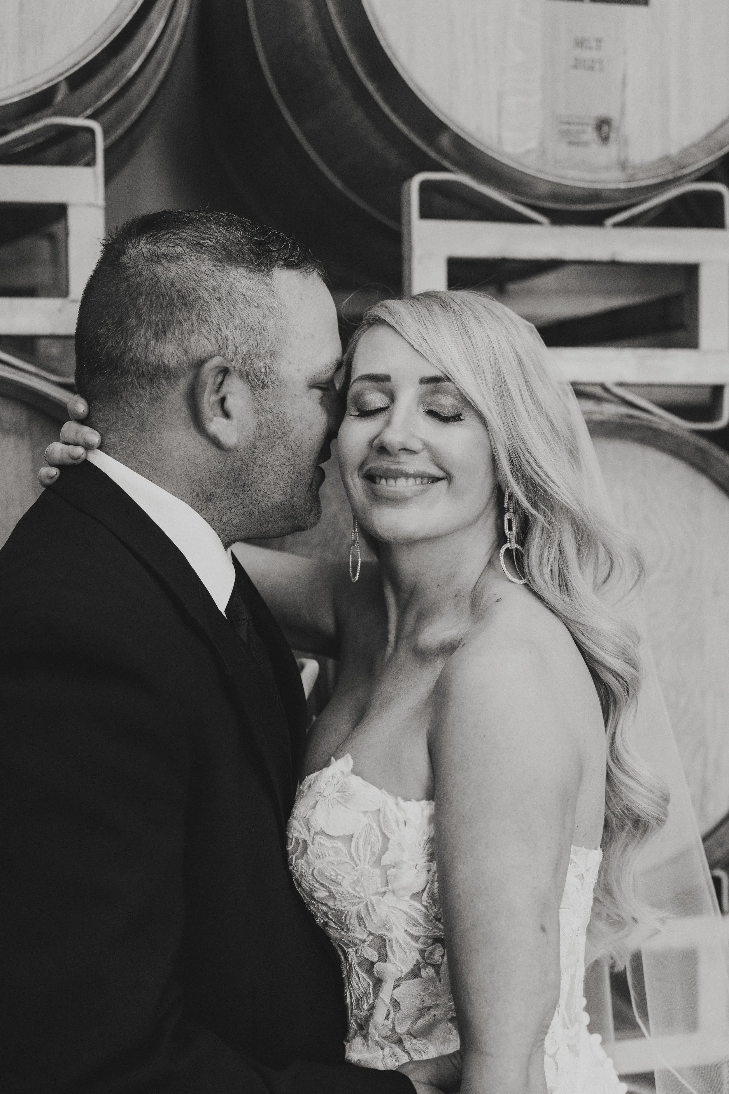 A black-and-white photo of a couple, with the man about to kiss the woman's forehead. The woman is smiling softly with her eyes closed, wearing earrings and a strapless lace dress. The man is wearing a tuxedo. They are standing in front of a backgrou