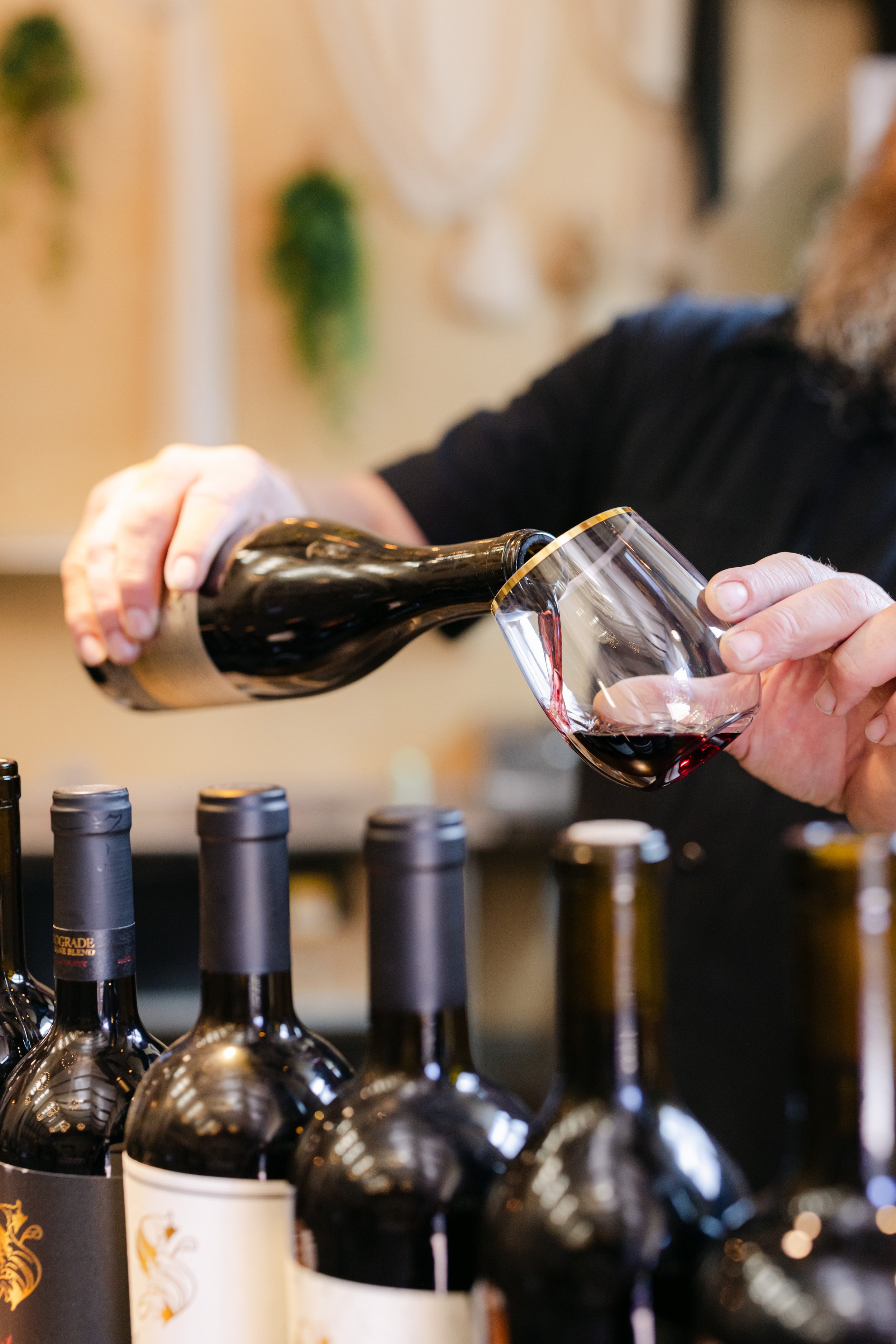 A person pouring red wine into a glass at a wine tasting event, with several wine bottles lined up in the foreground.