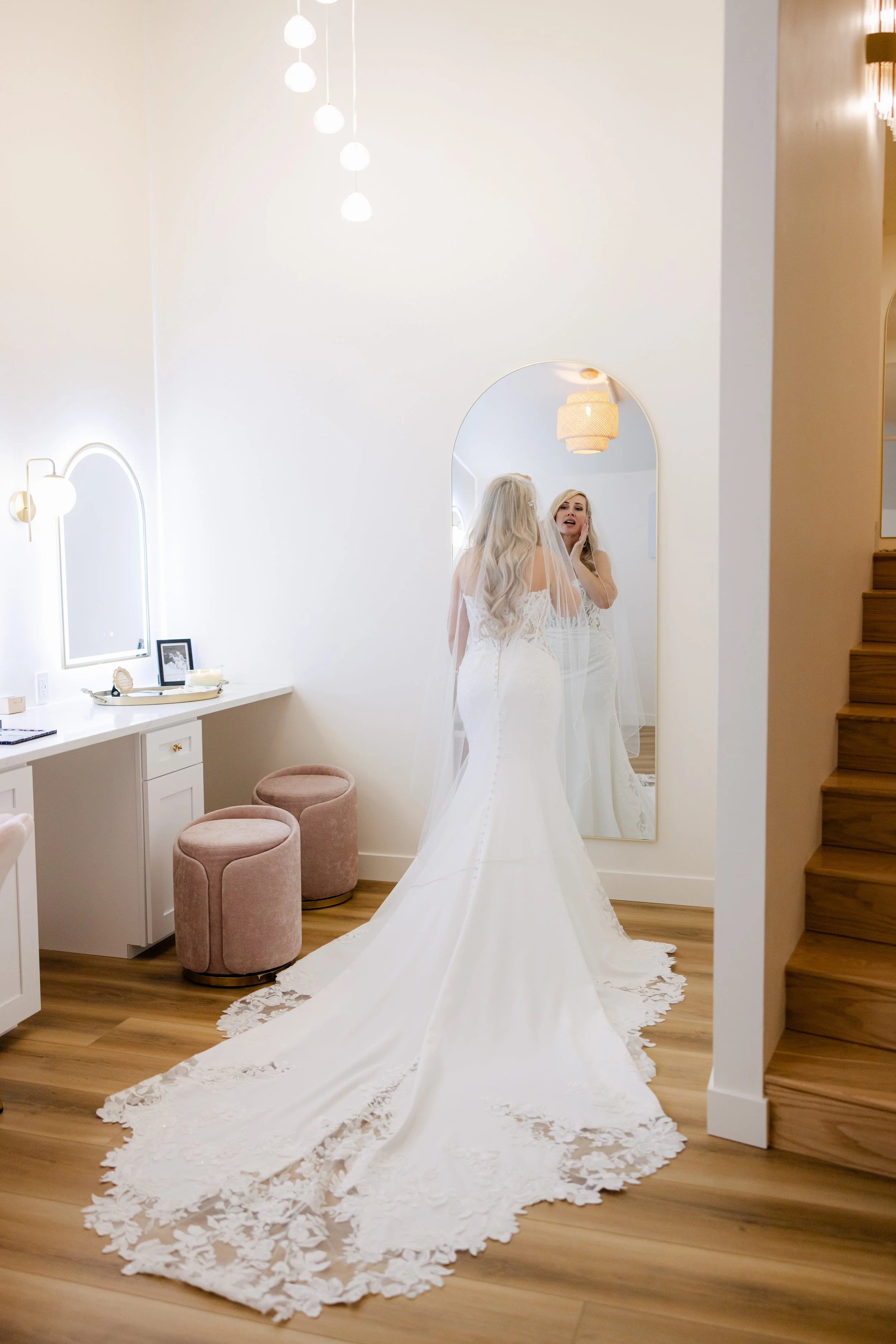 A bride in a wedding dress looking in a mirror, with a long train and veil in a bright, modern room.