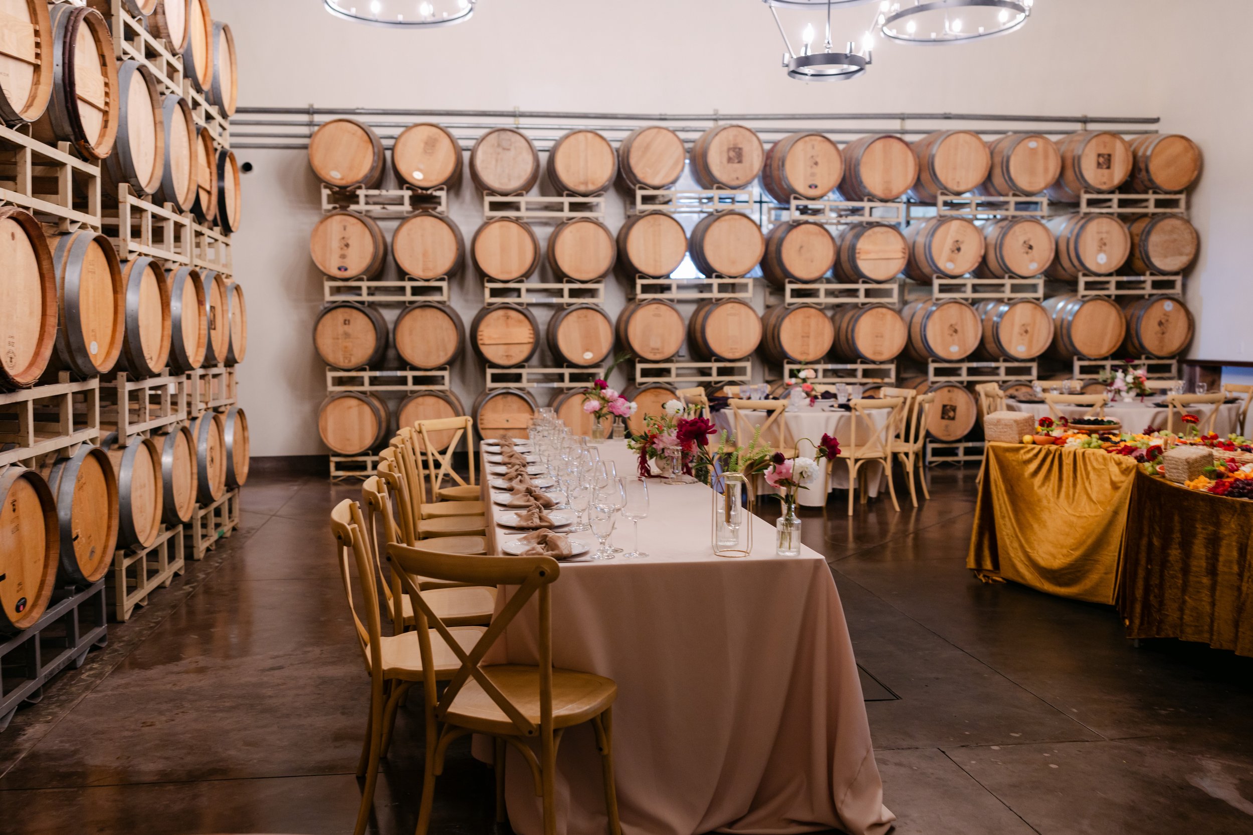 A wine cellar with stacked wooden barrels on metal racks. In the foreground, there is a long dining table with a beige tablecloth, set with glasses, napkins, and condiments. The table is decorated with flowers. To the right, there is a table covered 