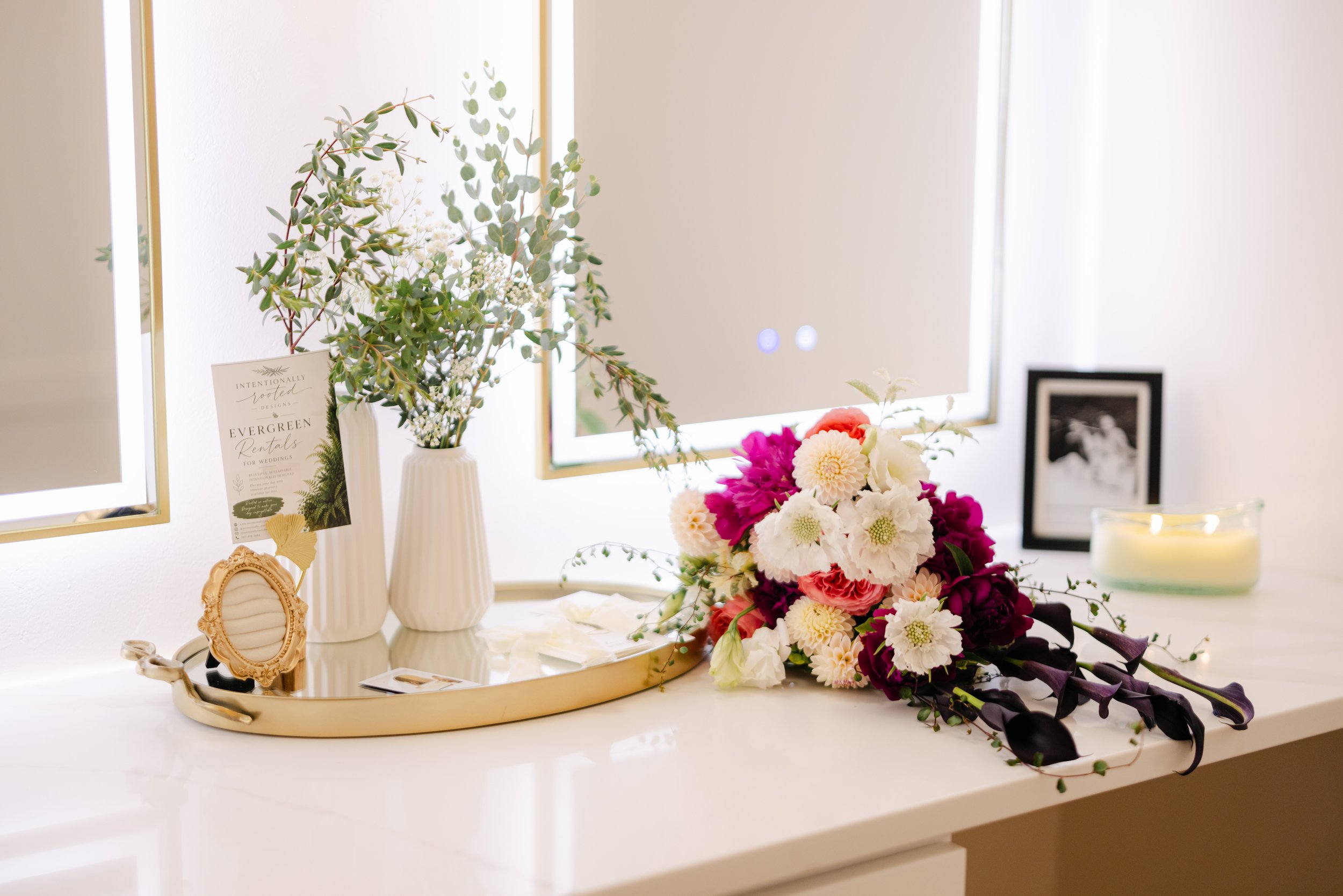 A white table decorated with a tray holding a small, ornate picture frame, a yellow hairband, and feminine accessories. Behind it, a bouquet of pink, white, and purple flowers is displayed, along with a white striped vase holding tall, leafy greenery