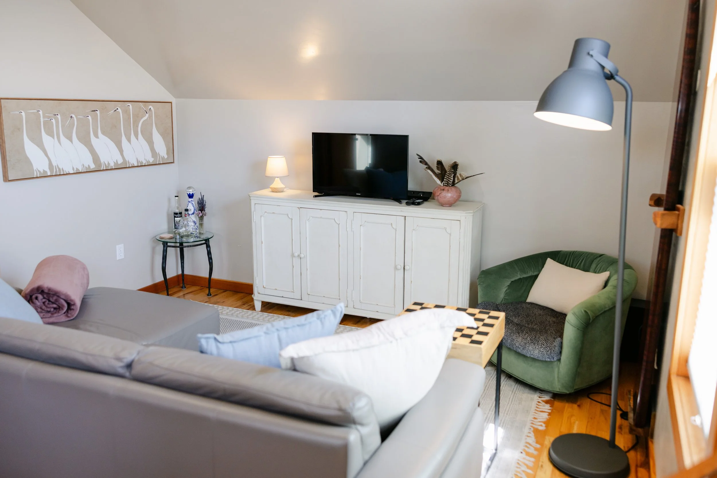 A living room with a white dresser, modern television, a table lamp, decorative vase with feathers, a green armchair, a wall art of white cranes, and a wall-mounted lamp.